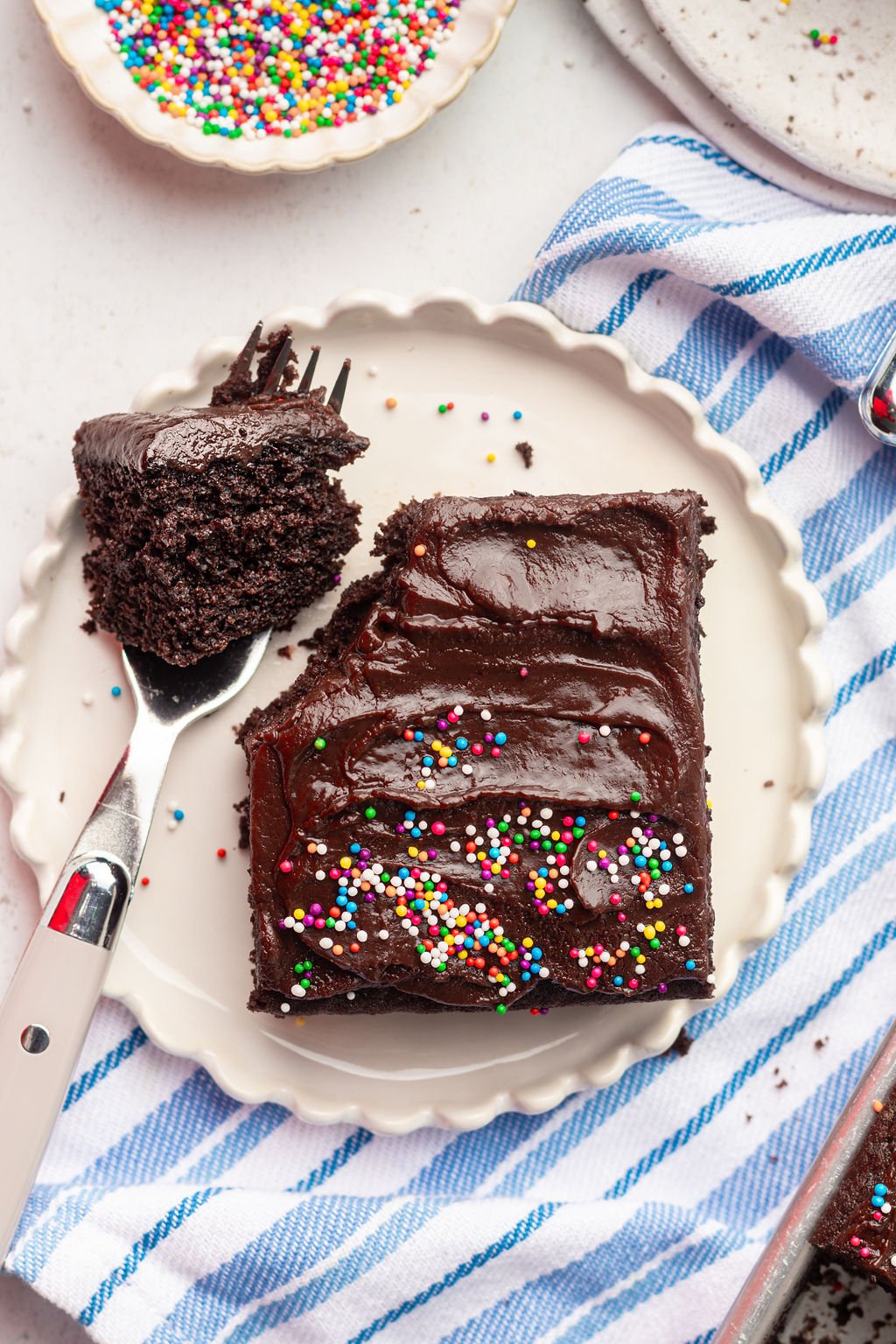 Chocolate cake with chocolate frosting and multi colored sprinkles on a white plate with a fork holding a bite of the square.