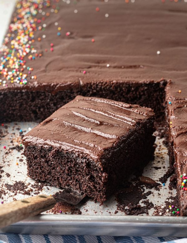 Chocolate sheet cake with multi colored sprinkles on a half sheet pan with one slice removed with an offset spatula.