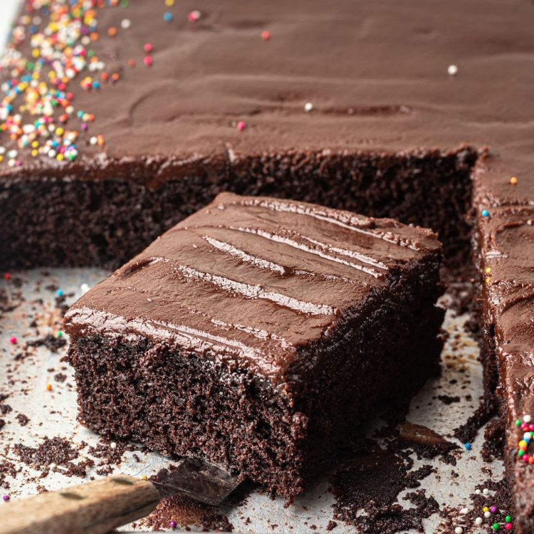 Chocolate sheet cake with multi colored sprinkles on a half sheet pan with one slice removed with an offset spatula.