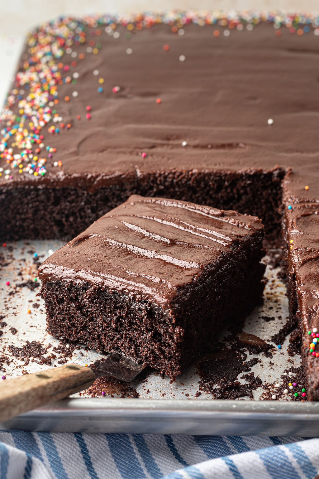 Chocolate sheet cake with multi colored sprinkles on a half sheet pan with one slice removed with an offset spatula.