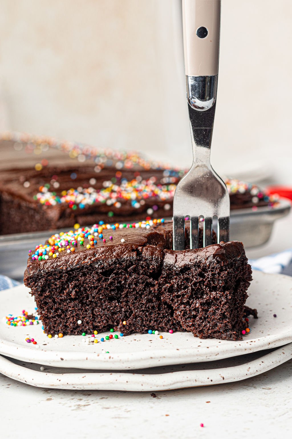 Square piece of chocolate cake with chocolate frosting and multi color sprinkles on a white plate with a fork stuck in the piece.