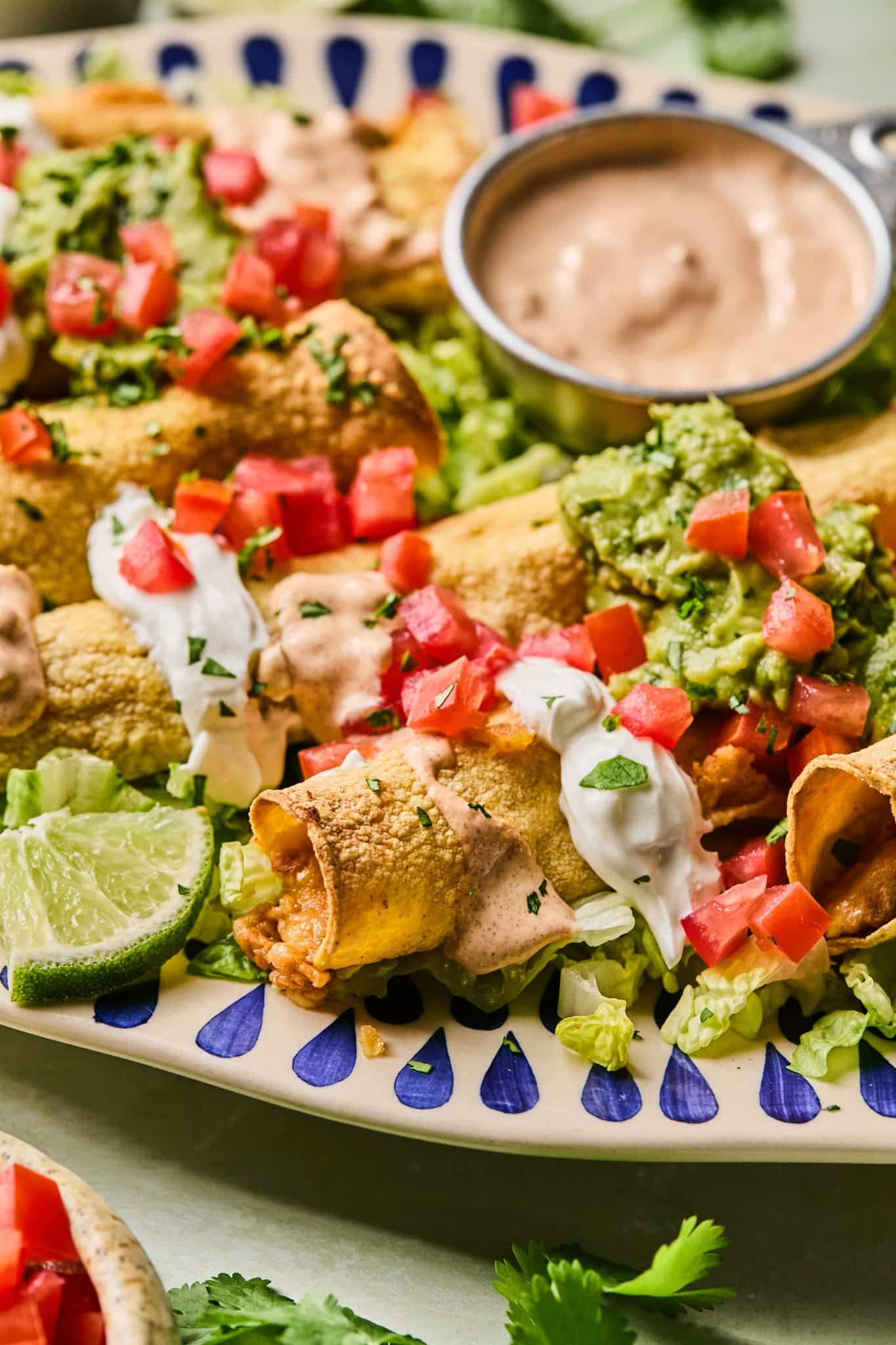 taquitos on a blue and white plate garnished with guacamole, sour cream, tomatoes and sauce. 