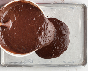 White mixing bowl with chocolate cake batter being poured onto a silver half sheet pan.