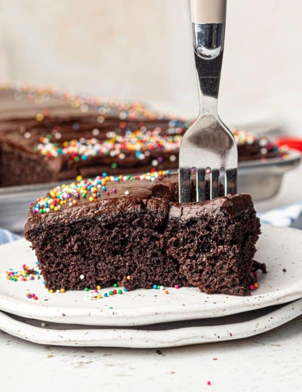 Square piece of chocolate cake with chocolate frosting and multi color sprinkles on a white plate with a fork stuck in the piece.