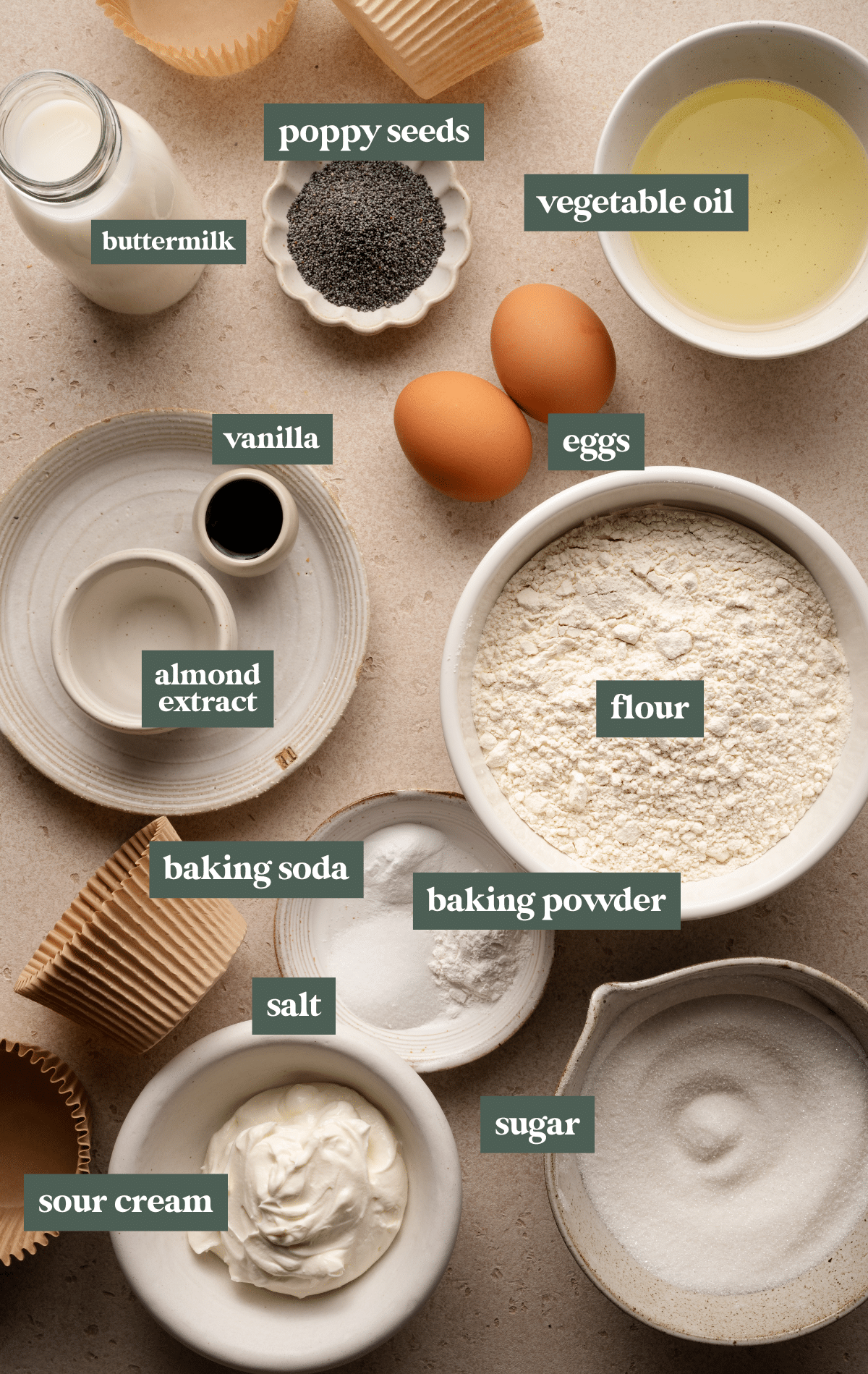 A flat-lay image of baking ingredients for almond poppy seed muffins, including labeled bowls of flour, sugar, poppy seeds, baking powder, baking soda, salt, sour cream, buttermilk, vegetable oil, vanilla extract, almond extract, and eggs. Muffin liners are also placed around the ingredients.