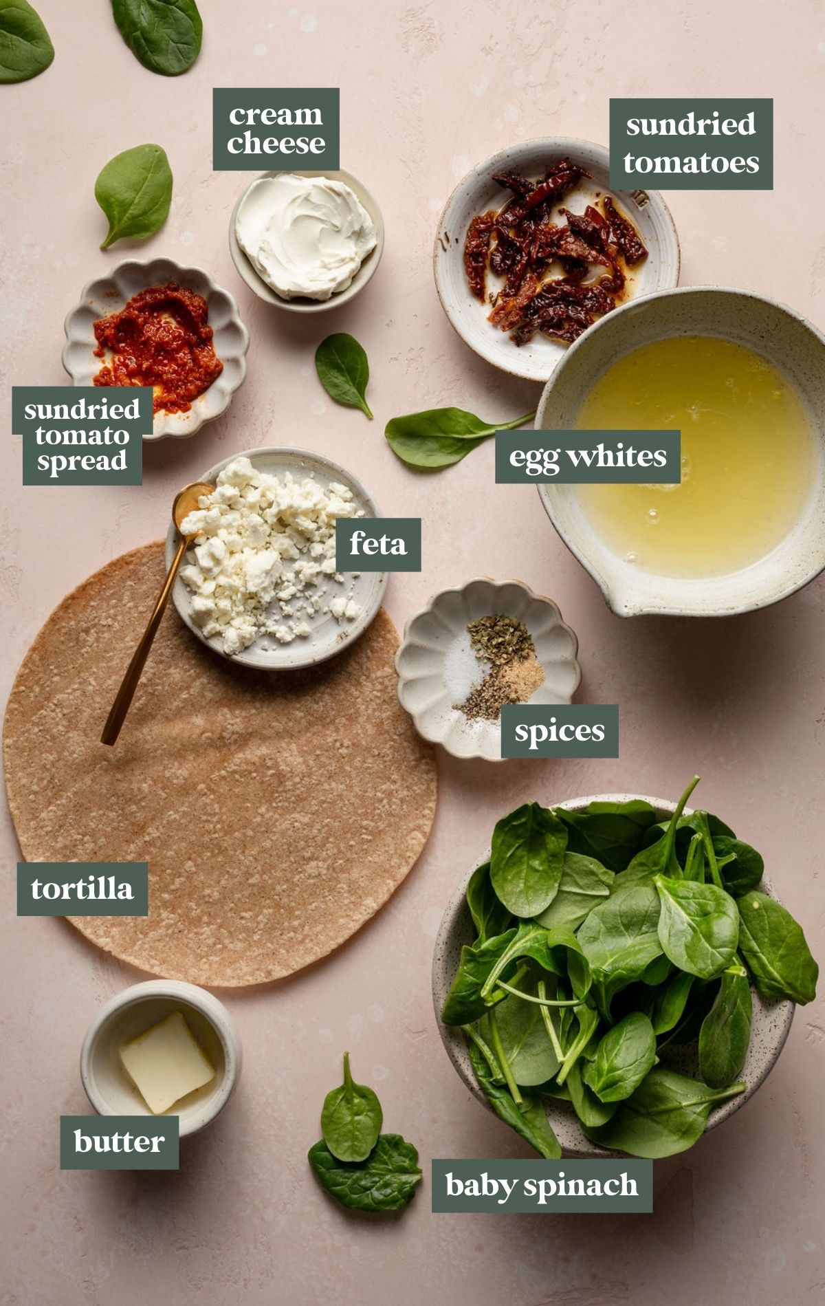 Flat lay of ingredients for a homemade Starbucks-style spinach and feta wrap, including a whole wheat tortilla, fresh spinach, egg whites, feta cheese, sun-dried tomatoes, spices, butter, and cream cheese spread, arranged on a soft pink background.