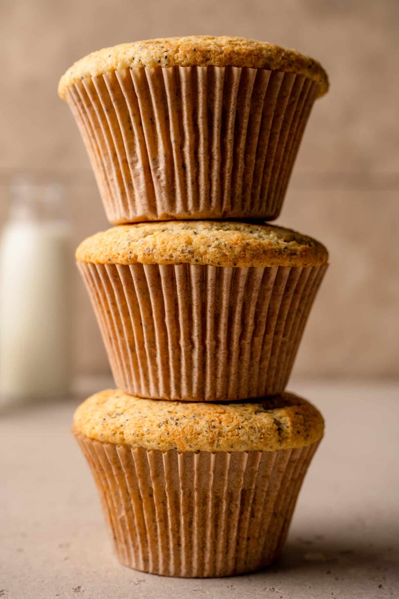  A stack of three golden-brown almond poppy seed muffins with a soft, fluffy texture, placed on a neutral surface. The muffins have a slightly domed top and visible poppy seeds throughout. A blurred milk bottle is visible in the background, adding a cozy, homemade feel.