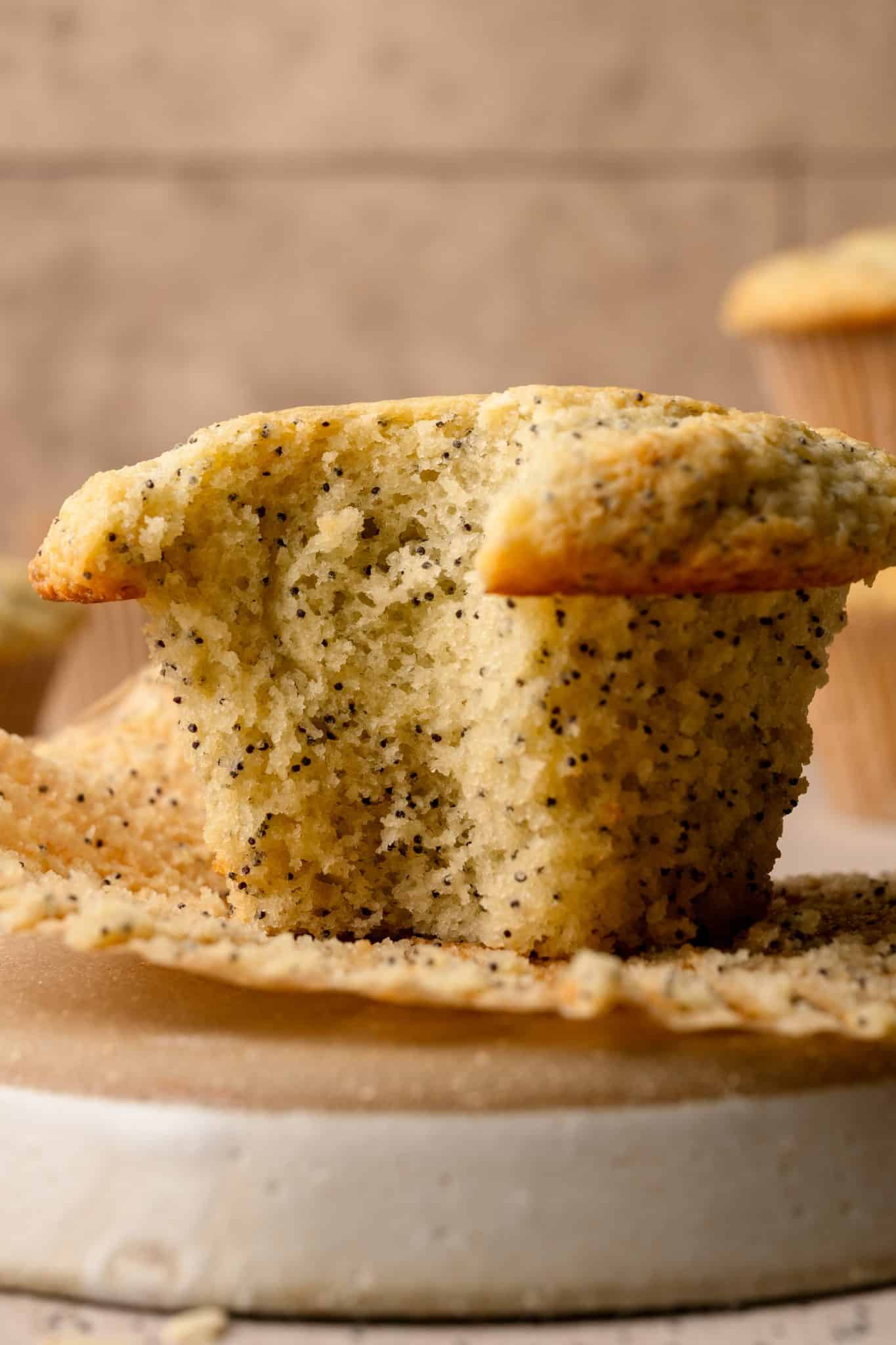 A close-up of a freshly baked almond poppy seed muffin, torn in half to reveal its soft, fluffy interior with evenly distributed poppy seeds. The muffin sits on an unwrapped liner with crumbs scattered around, highlighting its moist texture. A blurred background with additional muffins adds depth to the image.