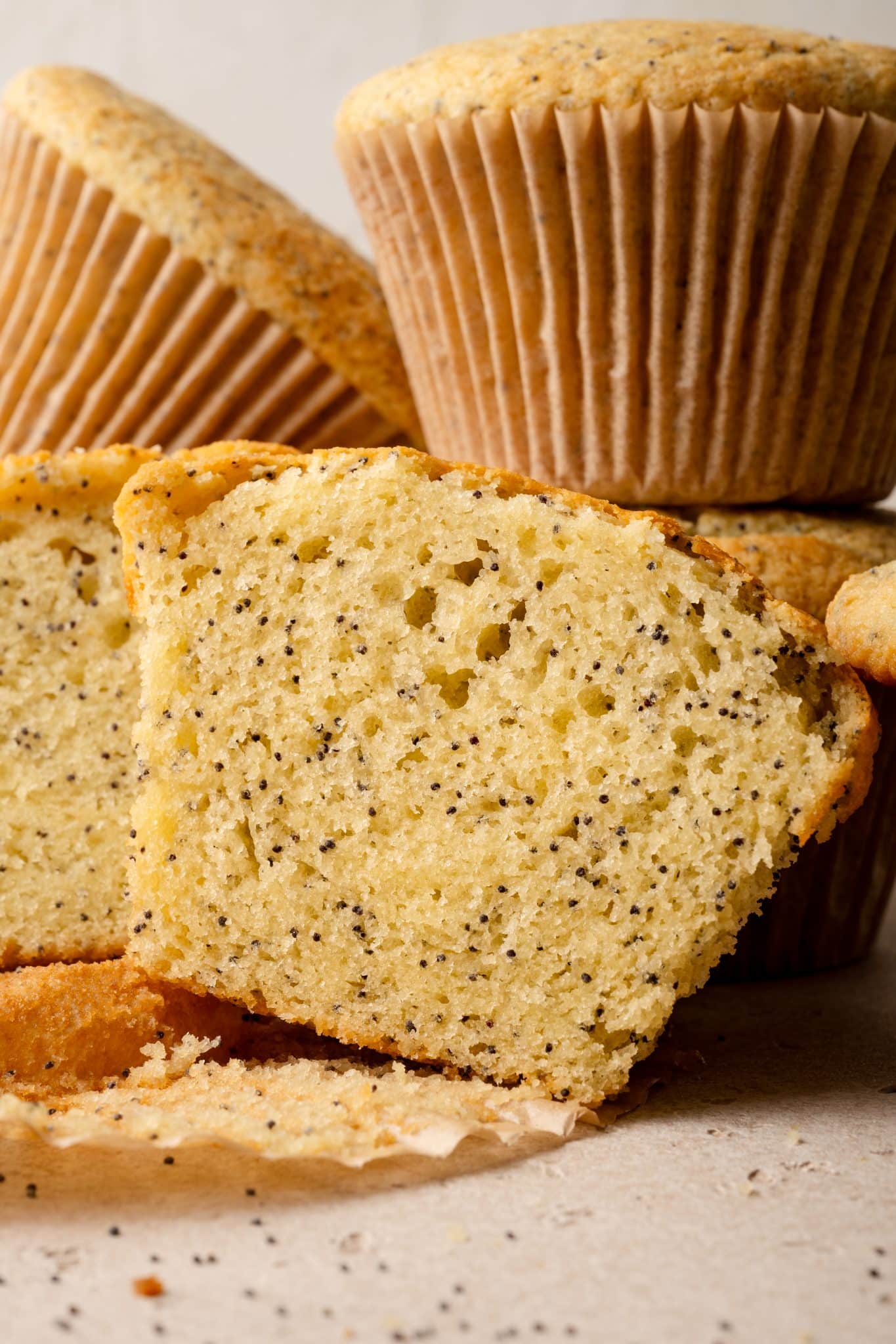 A close-up image of almond poppy seed muffins, with one muffin cut in half to reveal its soft, fluffy interior speckled with tiny black poppy seeds. The muffins have a golden-brown exterior and are stacked in the background, with crumbs and poppy seeds scattered on the surface.