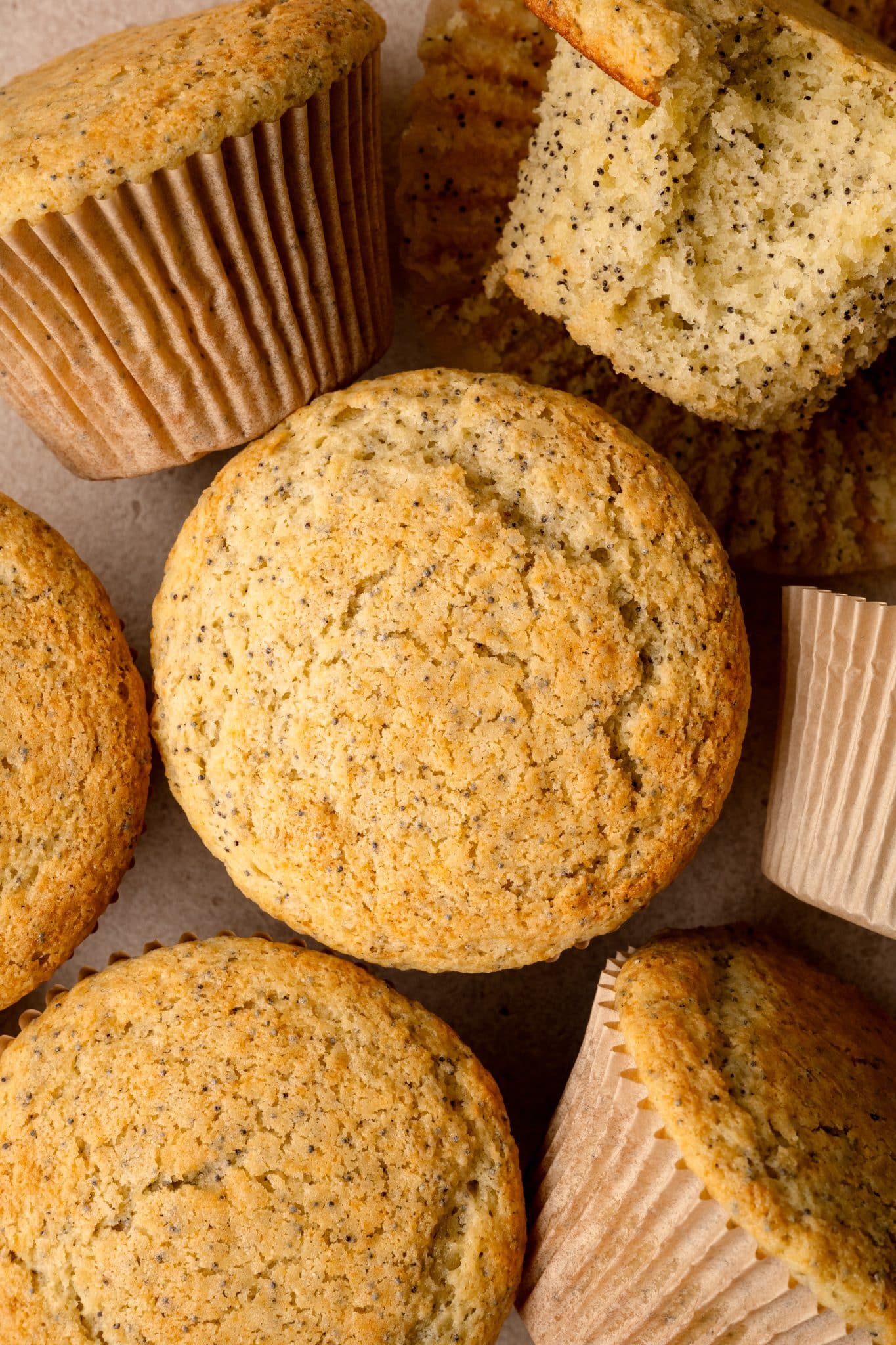 A close-up image of freshly baked almond poppy seed muffins. Some muffins are still in their paper liners, while others have been unwrapped, revealing a soft, fluffy interior with visible poppy seeds. The muffins have a golden-brown top and a textured surface.