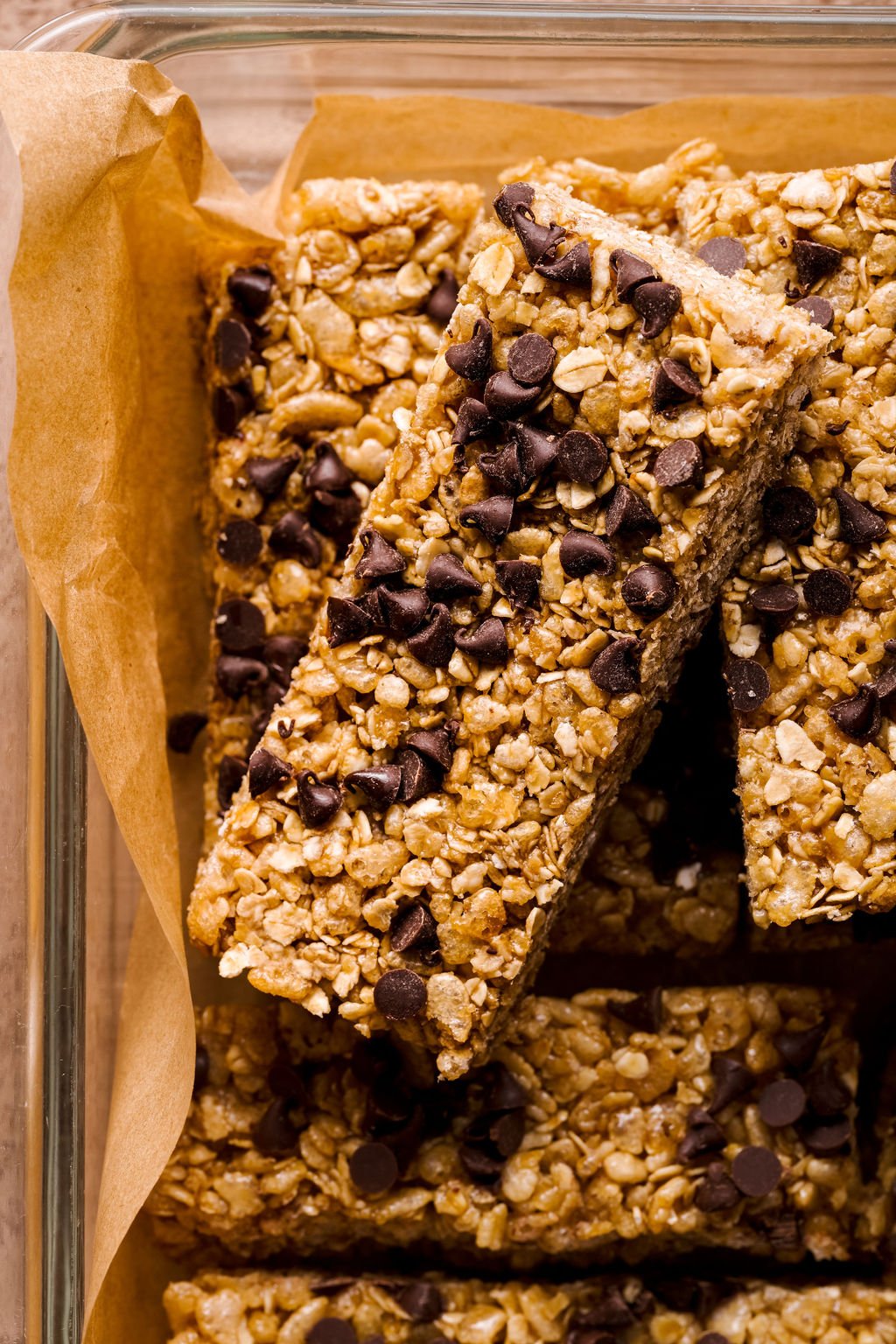 a close up image of a granola bar on parchment paper in a glass dish with mini chocolate chips on top. 