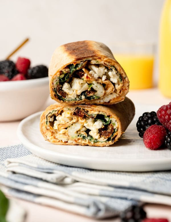 A close-up shot of a breakfast wrap cut in half and stacked on a white plate. The wrap has a golden-brown toasted tortilla and is filled with egg whites, spinach, sun-dried tomatoes, and crumbled feta cheese. The filling is slightly spilling out, revealing a mix of textures and colors. In the background, slightly out of focus, there is a bowl of fresh blackberries and raspberries, a glass of orange juice, and a folded napkin.