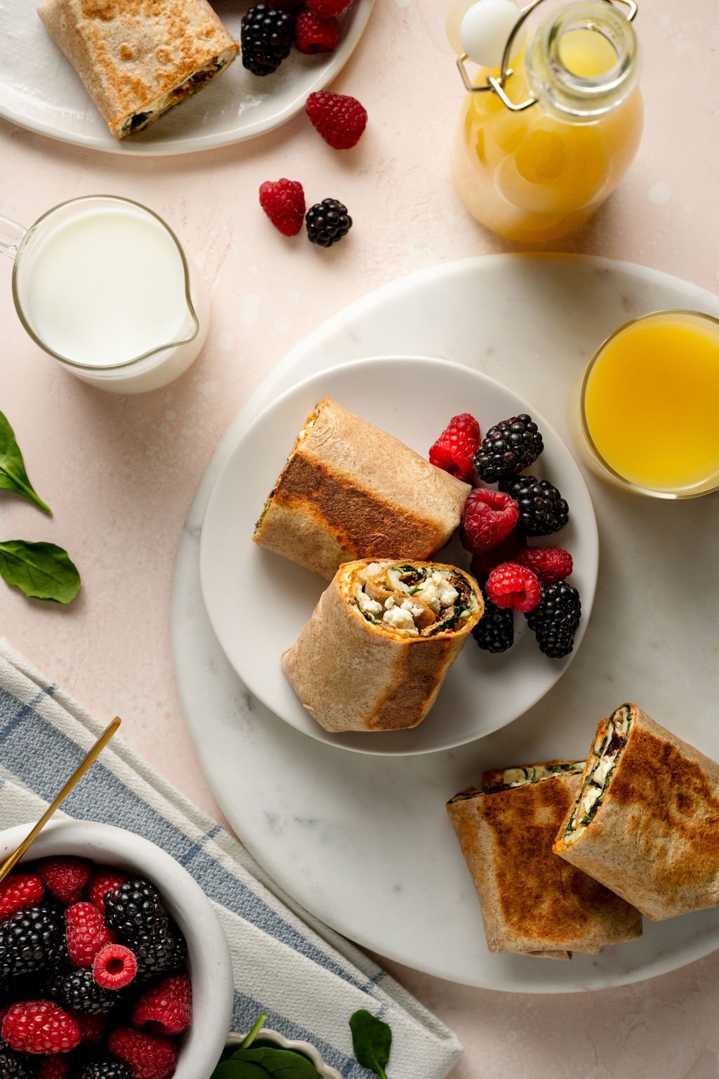 A flat-lay composition of a breakfast spread featuring multiple egg white breakfast wraps on white plates, accompanied by fresh berries (blackberries and raspberries), a glass of orange juice, and a small pitcher of milk. The scene is set on a light pink marble surface, with a folded blue-and-white checkered napkin and a golden spoon resting in a bowl of berries.