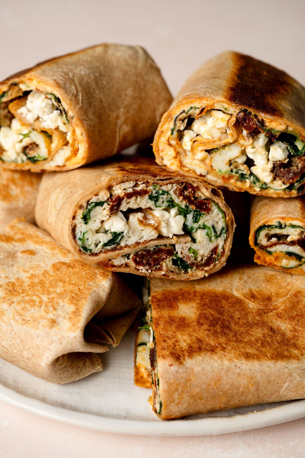 A close-up shot of several egg white breakfast wraps stacked together on a white plate. Some wraps are whole, while others are sliced open to reveal their filling of fluffy egg whites, wilted spinach, sun-dried tomatoes, and crumbled feta cheese. The tortillas have a golden-brown toasted exterior. The background is softly blurred, with a warm, inviting ambiance.