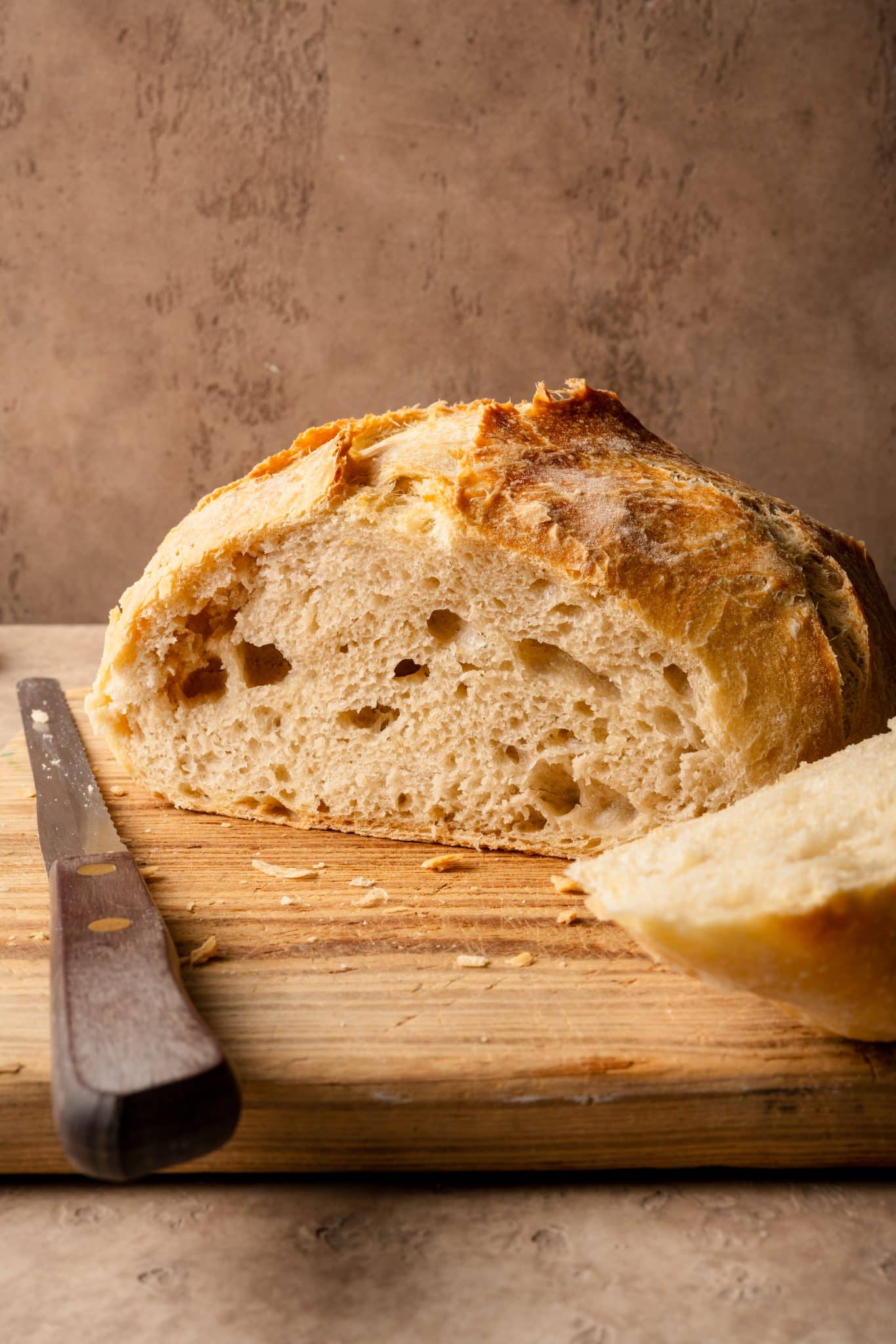 A crusty round loaf of no-knead bread with a soft, airy crumb, partially sliced on a wooden cutting board next to a knife.