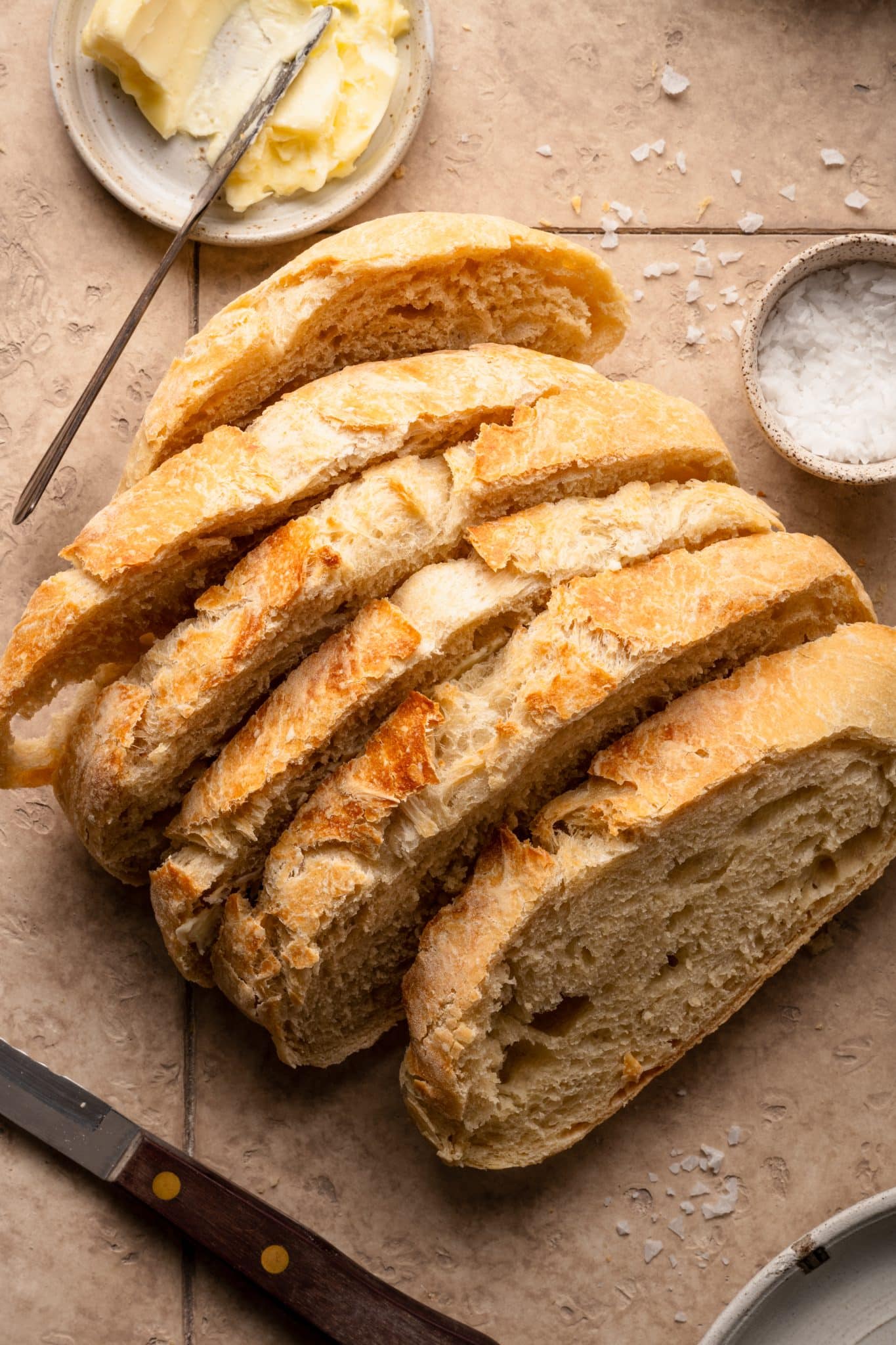 A sliced loaf of golden-brown, crusty no-knead bread on a light-colored surface, surrounded by a small dish of butter, a bowl of flaky salt, and a knife.