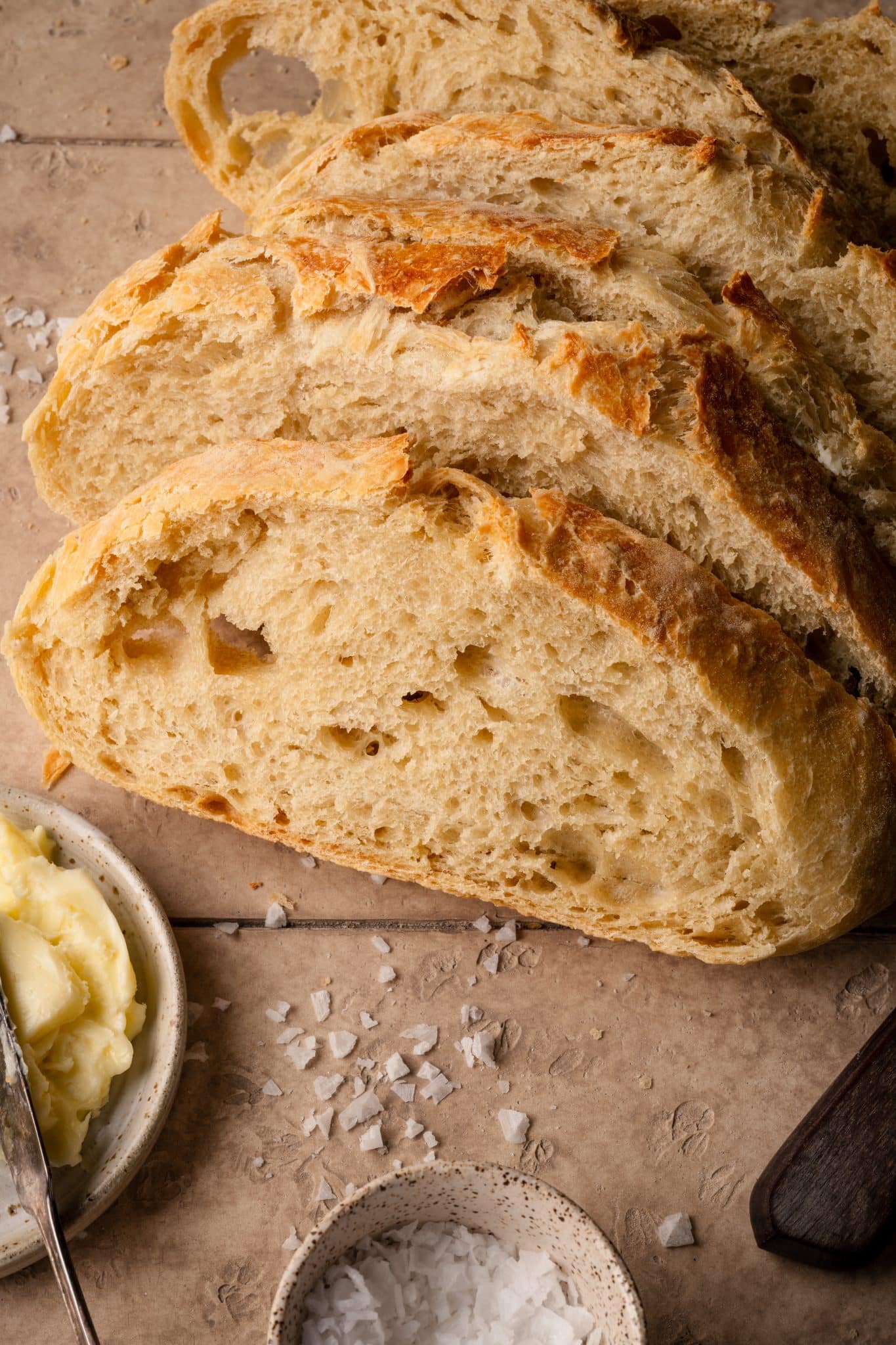 A close-up view of sliced, golden-brown no-knead bread with a soft, airy crumb, placed on a beige surface beside a dish of butter and a small bowl of flaky salt.