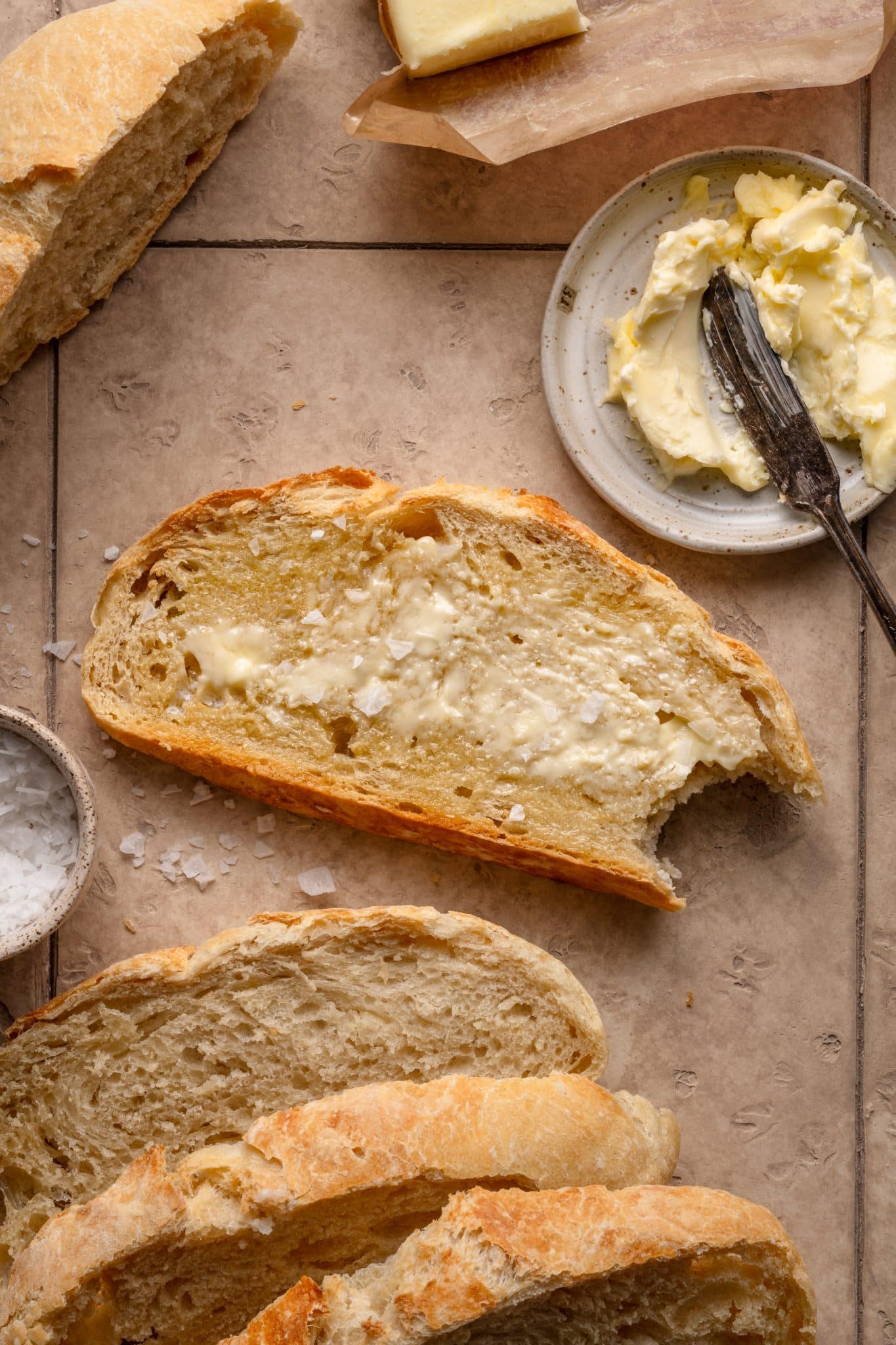 A slice of golden-brown no-knead bread spread with butter, surrounded by additional slices, a small butter dish, and a bowl of flaky salt on a tiled beige surface.