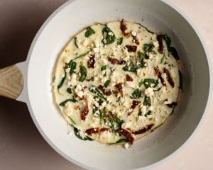 White frying pan with cooked egg whites, sundried tomatoes, spinach, and feta crumbled on top.