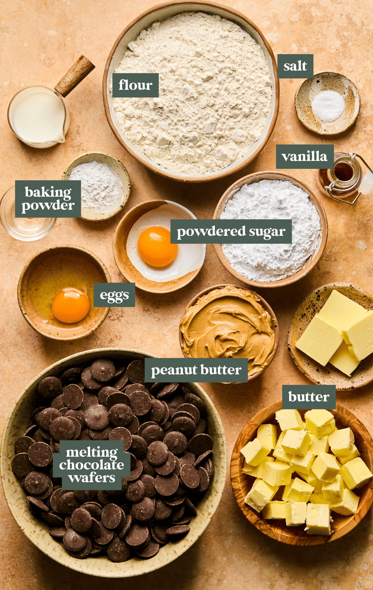 Bowls and measuring cups filled with flour, powdered sugar, peanut butter, butter, eggs, melting chocolate wafers, baking powder, salt, and vanilla, neatly arranged and labeled on a warm-toned surface.