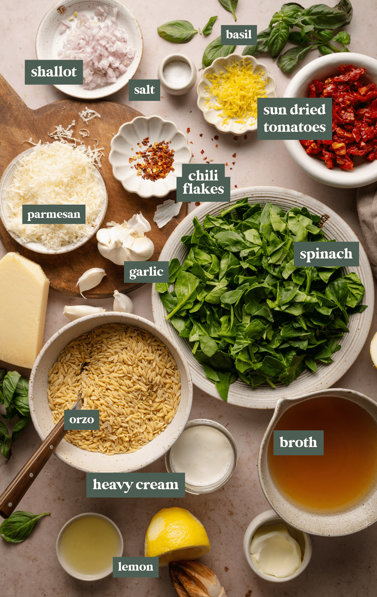 A flat-lay image showcasing the fresh ingredients for creamy sun-dried tomato and spinach orzo. The ingredients are neatly arranged and labeled, including orzo pasta, fresh spinach, sun-dried tomatoes, garlic, shallots, Parmesan cheese, basil, chili flakes, lemon zest, broth, heavy cream, and seasonings. The setup is on a neutral-toned surface with a rustic wooden board, highlighting the vibrant colors and textures of each ingredient.