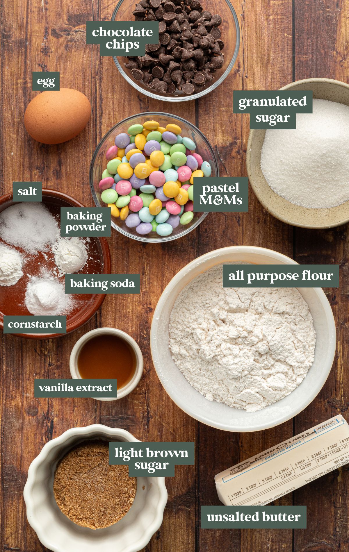 A top-down view of baking ingredients for spring-themed M&M cookies arranged on a wooden surface. The ingredients include a bowl of all-purpose flour, granulated sugar, light brown sugar, a stick of unsalted butter, an egg, vanilla extract, salt, baking powder, baking soda, cornstarch, chocolate chips, and pastel-colored M&M candies. Each ingredient is labeled with text overlays.