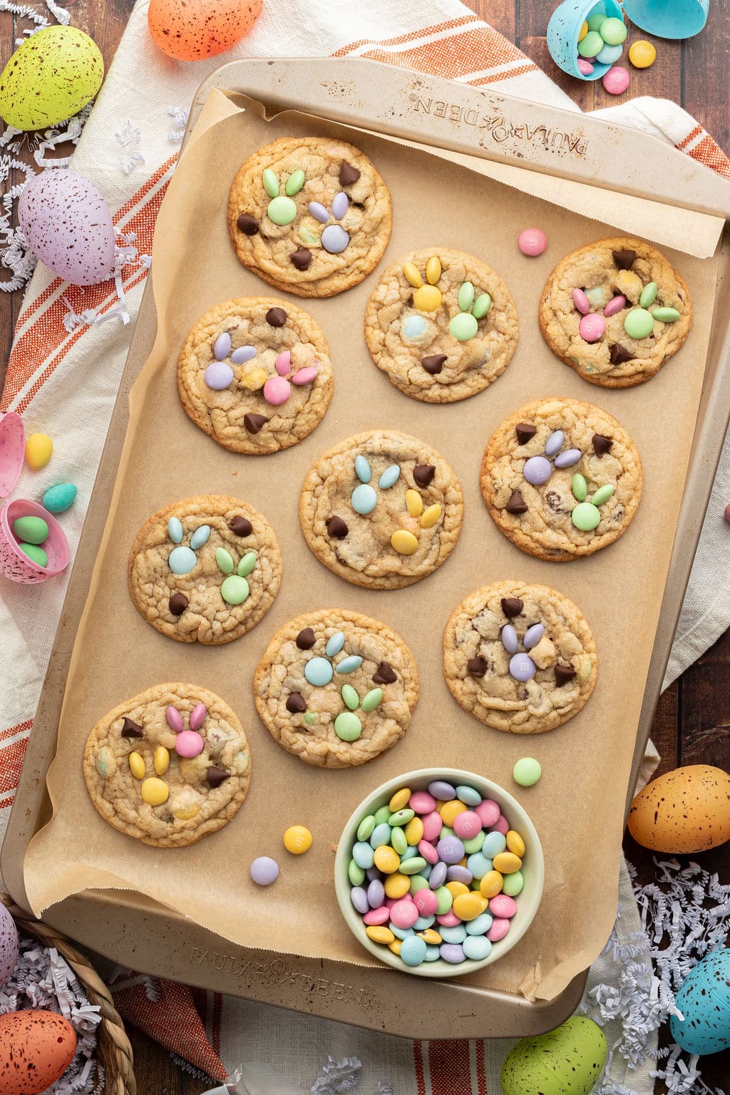 A baking sheet lined with parchment paper holding freshly baked M&M cookies decorated with pastel-colored candies arranged in a bunny shape. Surrounding the tray are speckled Easter eggs, a striped dish towel, and a bowl of extra M&Ms.