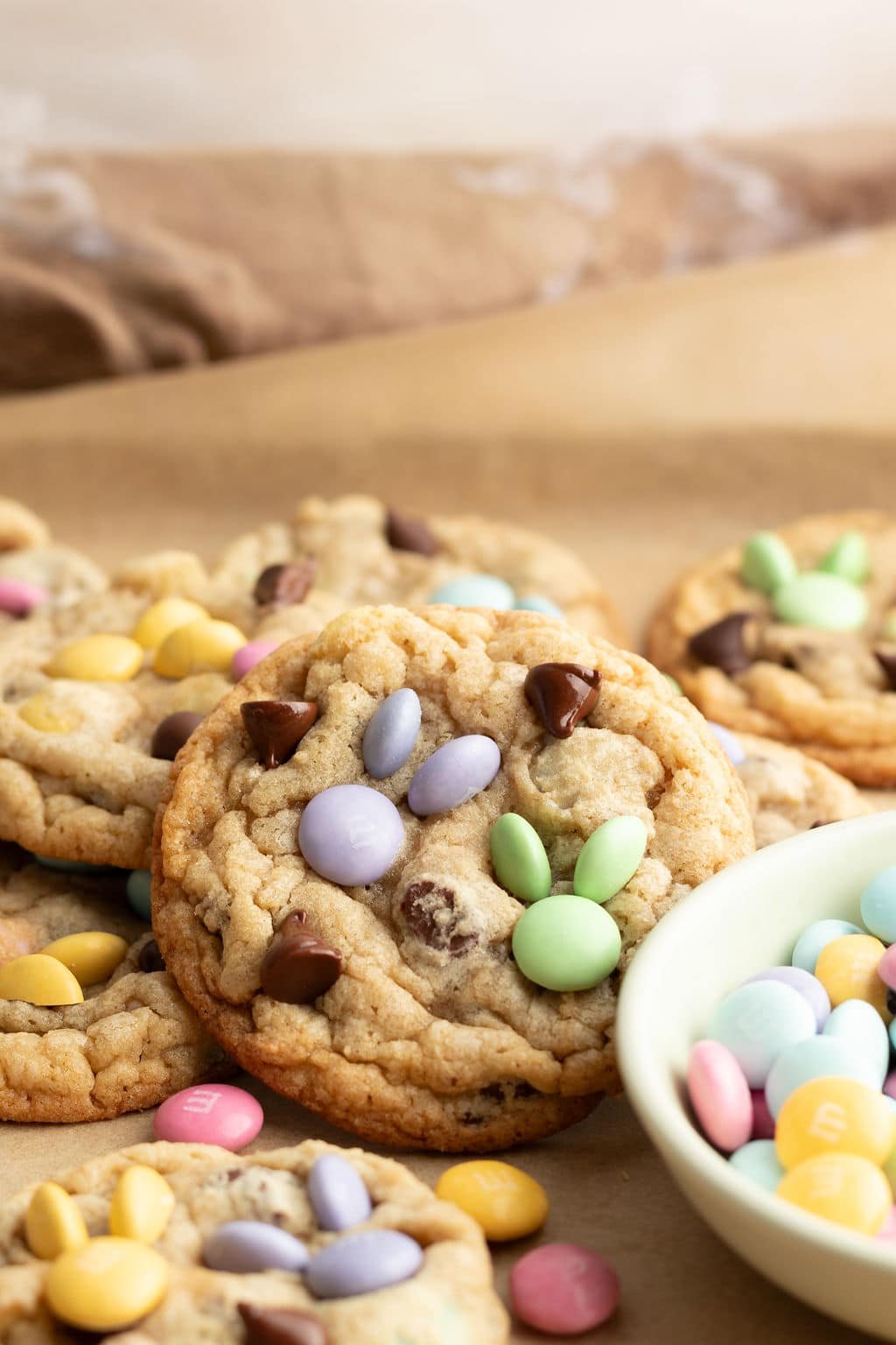 A close-up of a freshly baked cookie decorated with pastel M&Ms and chocolate chips, highlighting its chewy texture. Other cookies and a bowl of colorful M&Ms are visible in the background.