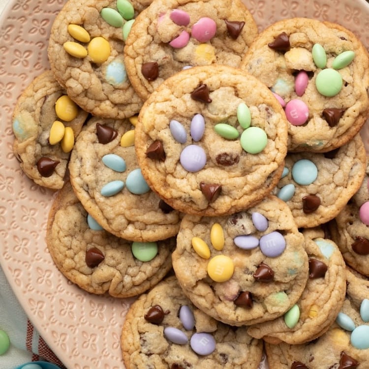 A decorative pink plate filled with spring-themed M&M cookies, featuring pastel-colored candies arranged in bunny designs. A bowl of extra M&Ms and festive Easter decorations surround the plate.