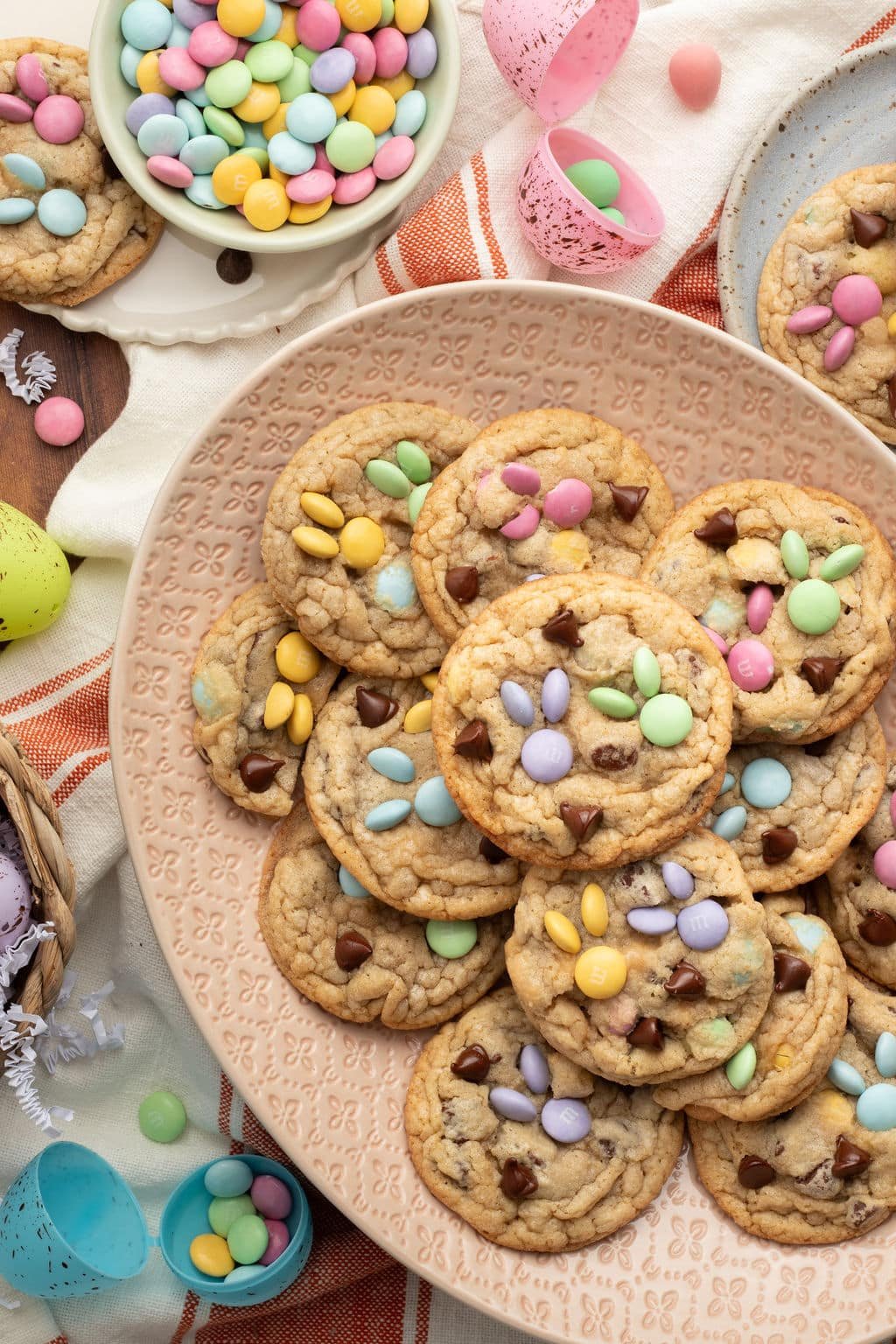 A decorative pink plate filled with spring-themed M&M cookies, featuring pastel-colored candies arranged in bunny designs. A bowl of extra M&Ms and festive Easter decorations surround the plate.