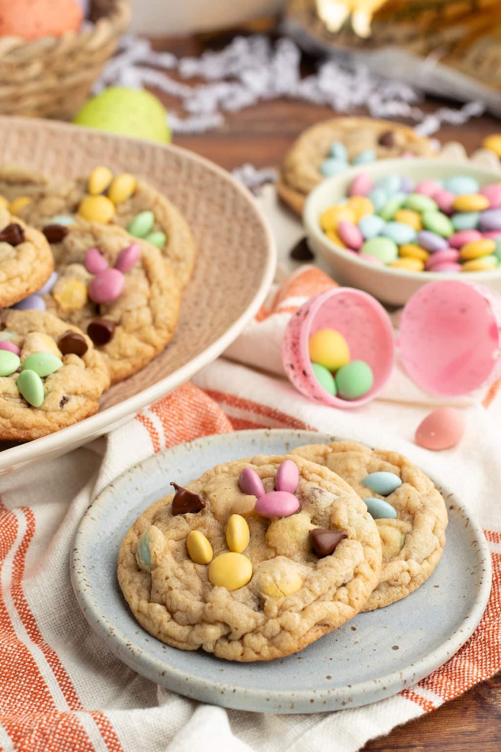 A small blue plate with two M&M cookies on a striped dish towel. One cookie is topped with pink and yellow candies, while the other features blue and green candies. The background includes a basket of cookies and pastel Easter decorations.