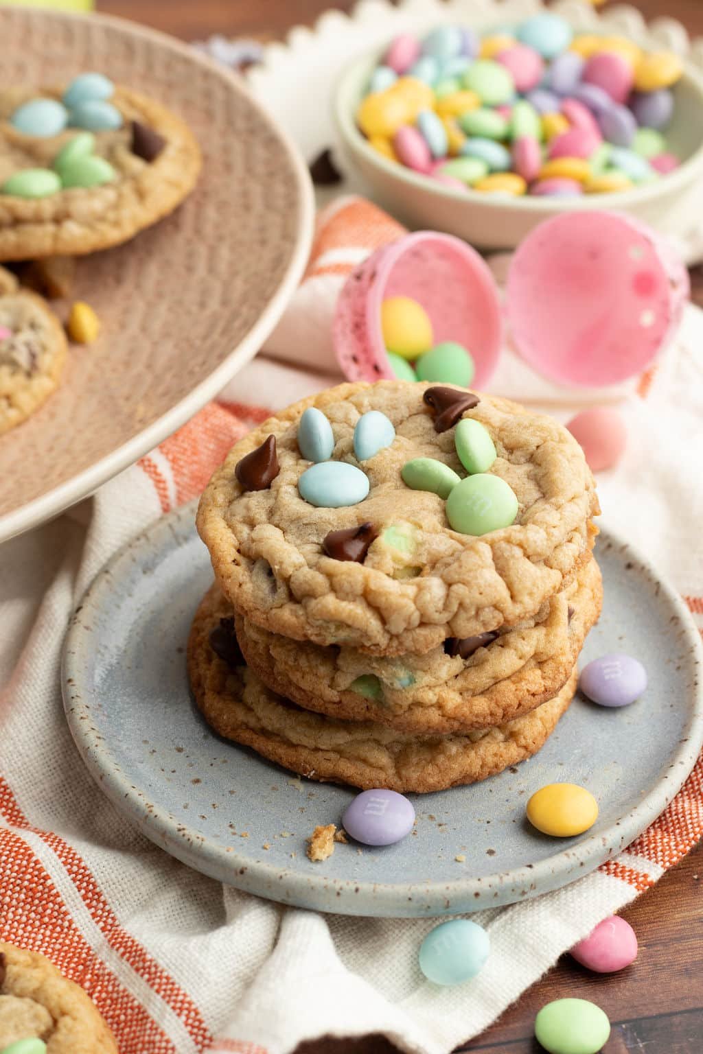 A stack of three chewy M&M cookies on a rustic blue plate, topped with pastel candies in the shape of bunnies and chocolate chips. Around the plate are scattered M&Ms and decorative Easter eggs.