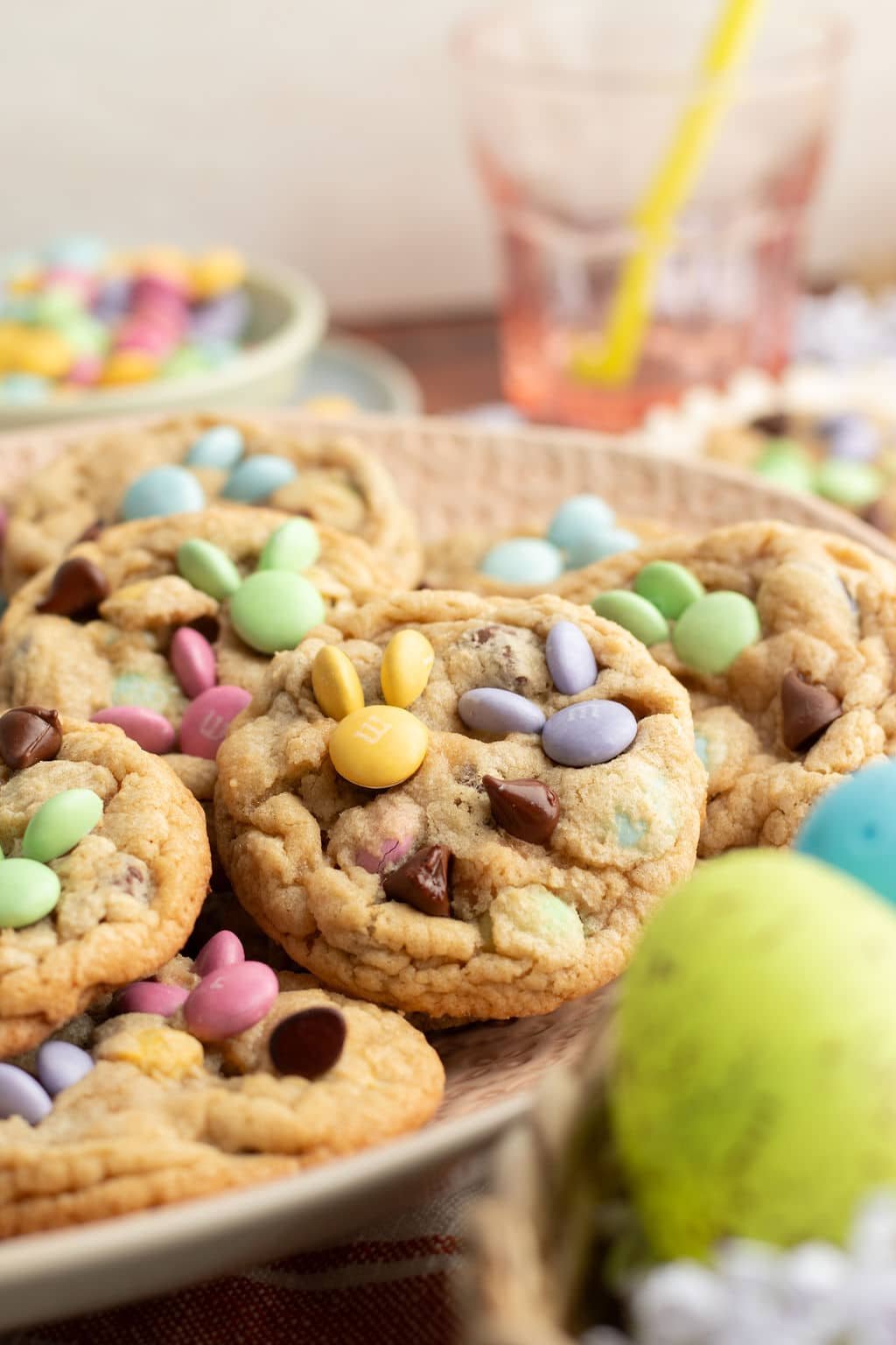 A close-up of a plate filled with freshly baked M&M cookies. The pastel-colored candies and chocolate chips are arranged in festive bunny shapes, with a blurred background of a drink and other spring decorations.