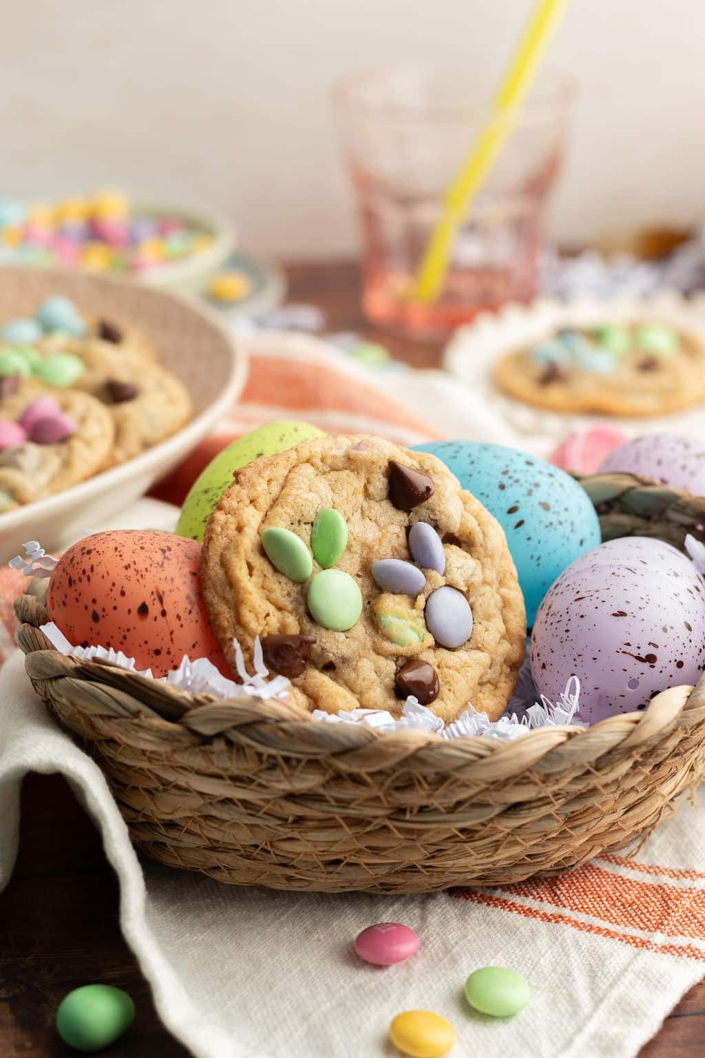 A woven Easter basket filled with speckled decorative eggs and a single M&M cookie decorated with pastel candies in the shape of a bunny. The background includes additional cookies and a spring-themed table setting.