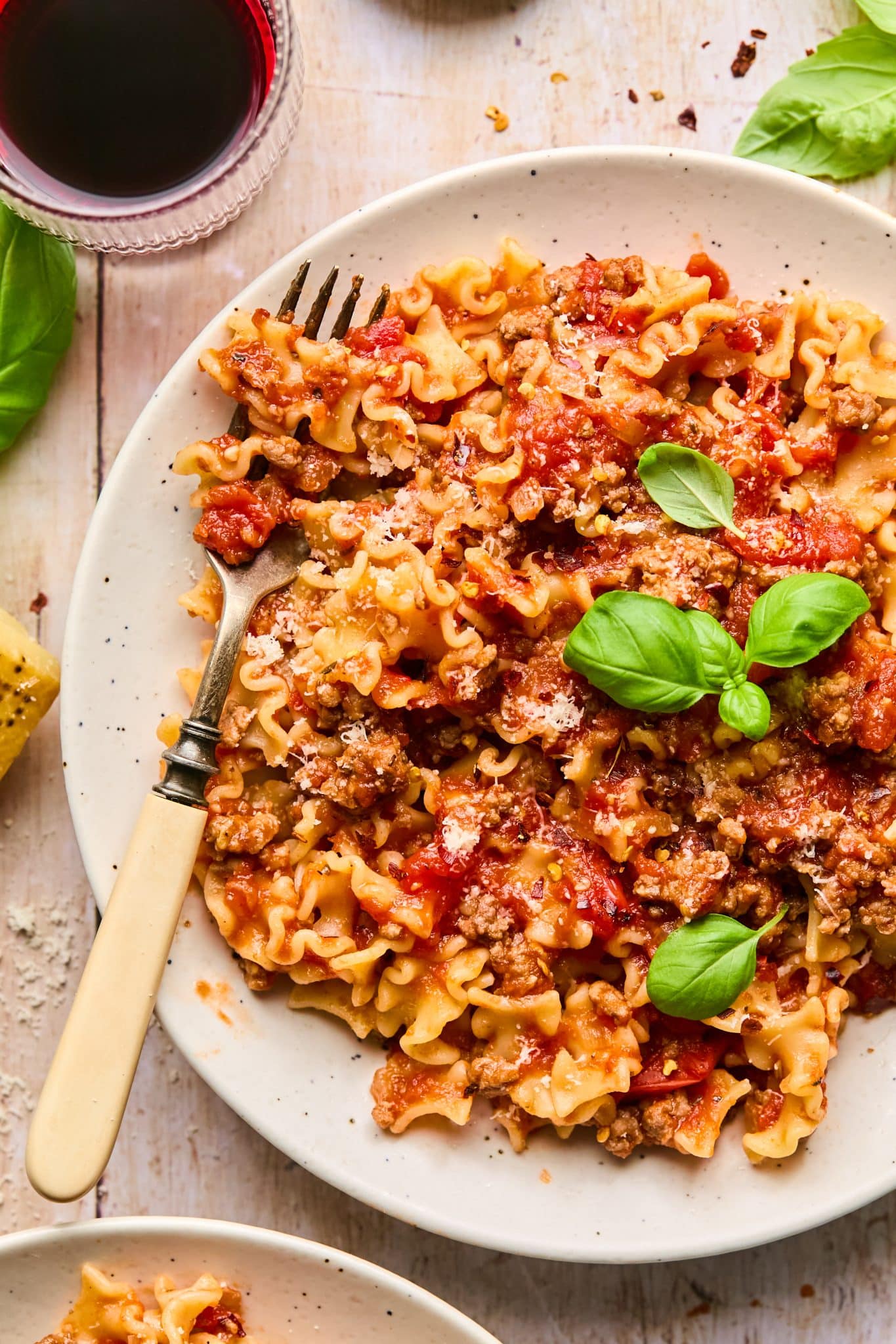 A top-down view of a plate of pasta with slow-cooked spaghetti sauce, garnished with fresh basil leaves and grated Parmesan cheese. A vintage fork is partially twirled into the pasta, and a glass of red wine sits nearby on a rustic wooden table, accompanied by scattered basil leaves and a block of Parmesan cheese.