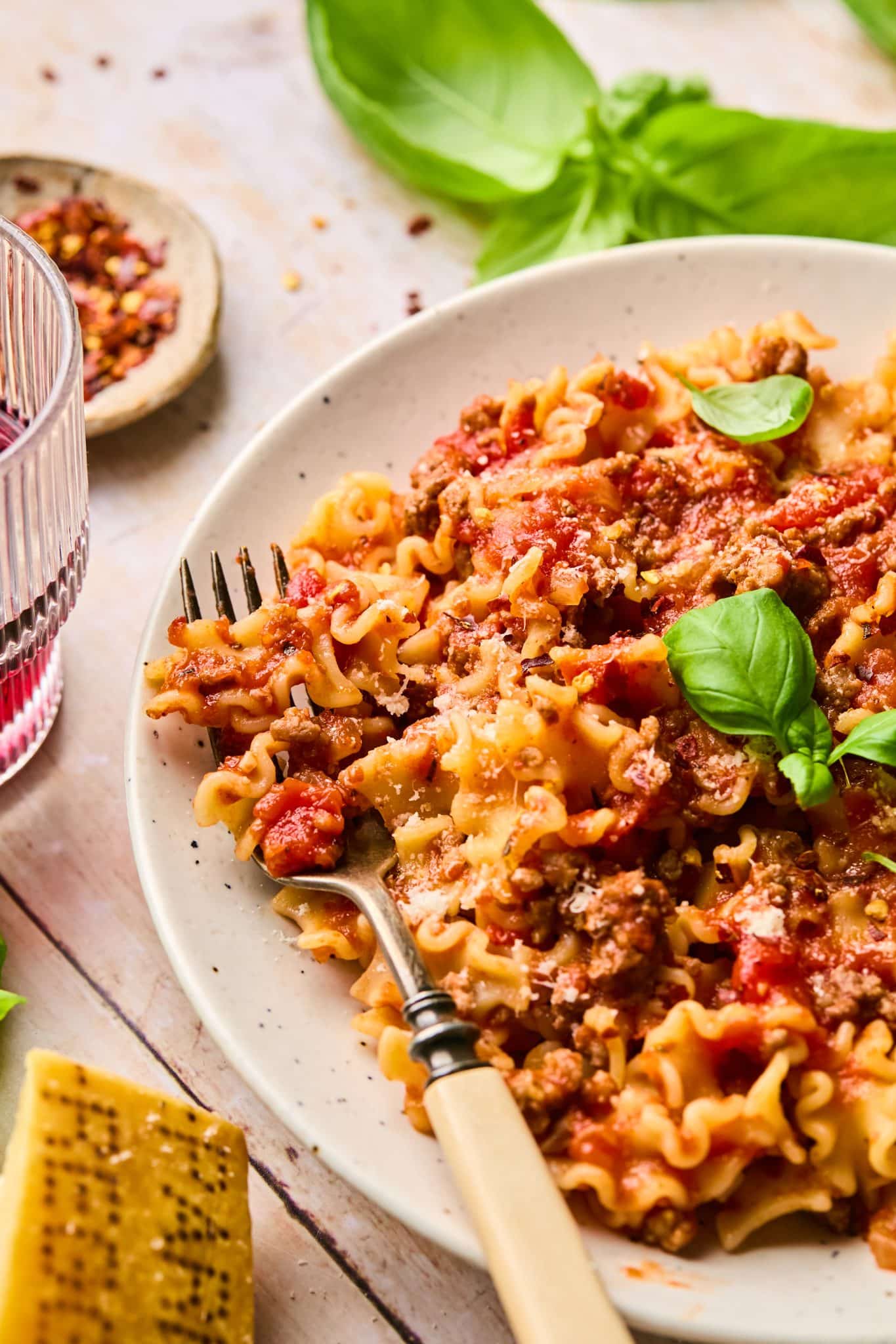 A close-up of a plate of pasta with slow-cooked spaghetti sauce, garnished with fresh basil leaves and grated Parmesan cheese. A vintage fork is twirled into the pasta, and a glass of red wine sits nearby. In the background, fresh basil leaves, crushed red pepper flakes, and a block of Parmesan cheese are visible on a rustic wooden table.