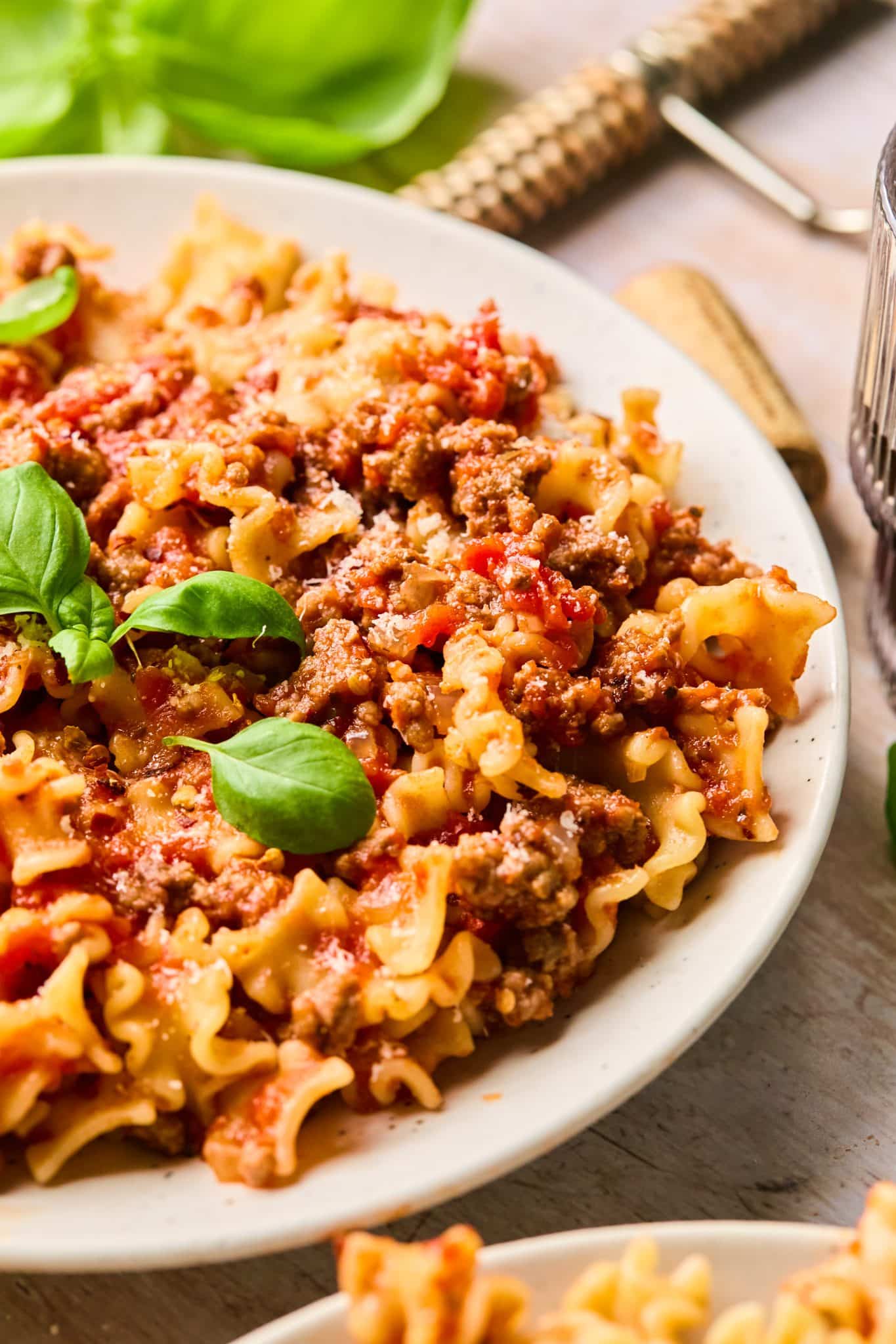 A close-up of a plate of pasta topped with slow-cooked spaghetti sauce, made with ground meat, tomatoes, and seasonings. The dish is garnished with fresh basil leaves and a sprinkle of grated Parmesan cheese. In the background, a grater, cork, and glass are visible, adding to the rustic and cozy dining setting.