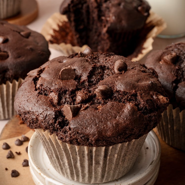 Image of double chocolate bakery-style muffin in paper liner on top of two mini serving plates and 4 muffins in the background.