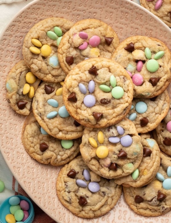 A decorative pink plate filled with spring-themed M&M cookies, featuring pastel-colored candies arranged in bunny designs. A bowl of extra M&Ms and festive Easter decorations surround the plate.