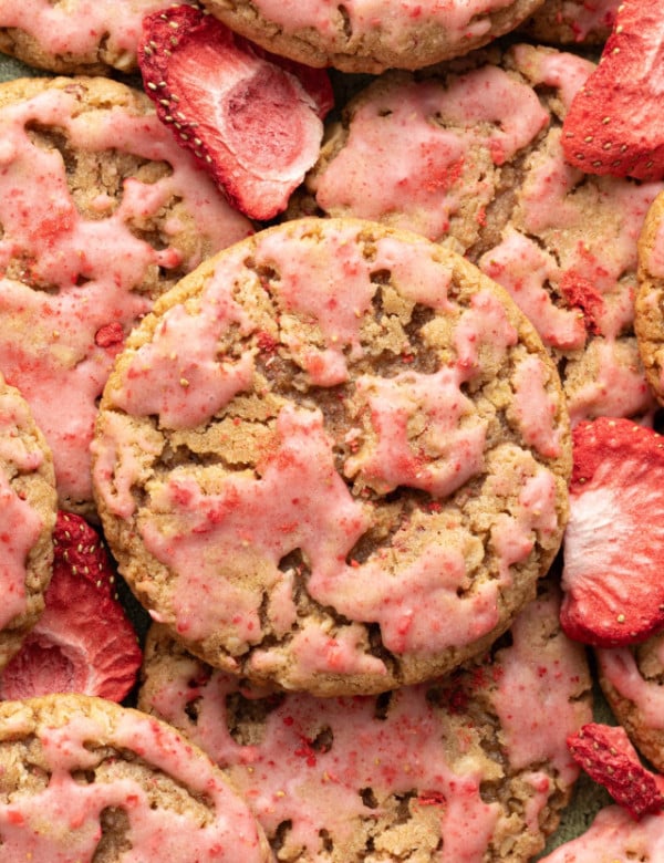 A close-up overhead shot of strawberry-glazed oatmeal cookies, showing their craggy texture and pink glaze. Freeze-dried strawberry slices are scattered among the cookies.