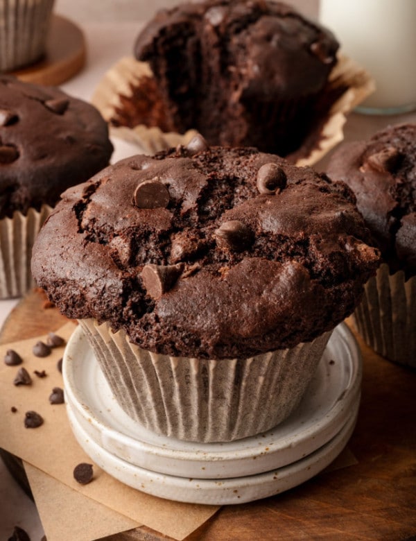 Image of double chocolate bakery-style muffin in paper liner on top of two mini serving plates and 4 muffins in the background.