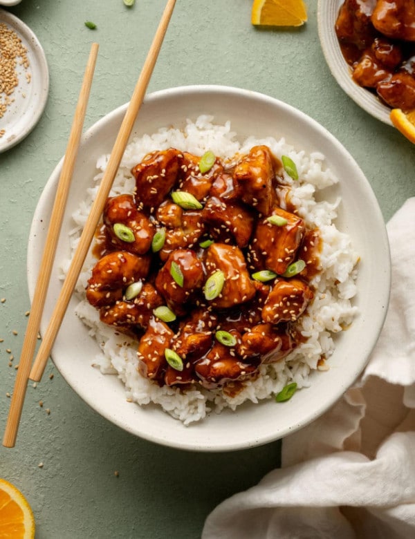 Plate of panda express orange chicken in bowl on top of white rice garnished with green onions sesame seeds with pair of chopsticks on the side.