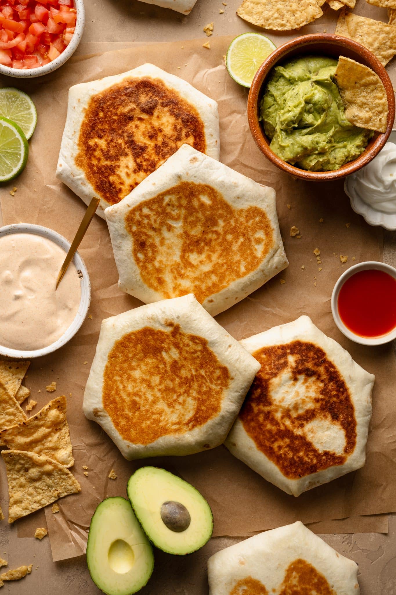 Golden brown homemade Crunchwrap Supremes arranged on parchment paper, with crisped tortillas folded into a hexagonal shape. Surrounding the wraps are fresh avocado halves, a bowl of guacamole with tortilla chips, a dish of sour cream, a small bowl of hot sauce, and a creamy dipping sauce with a spoon. Lime slices and diced tomatoes add fresh color to the composition.