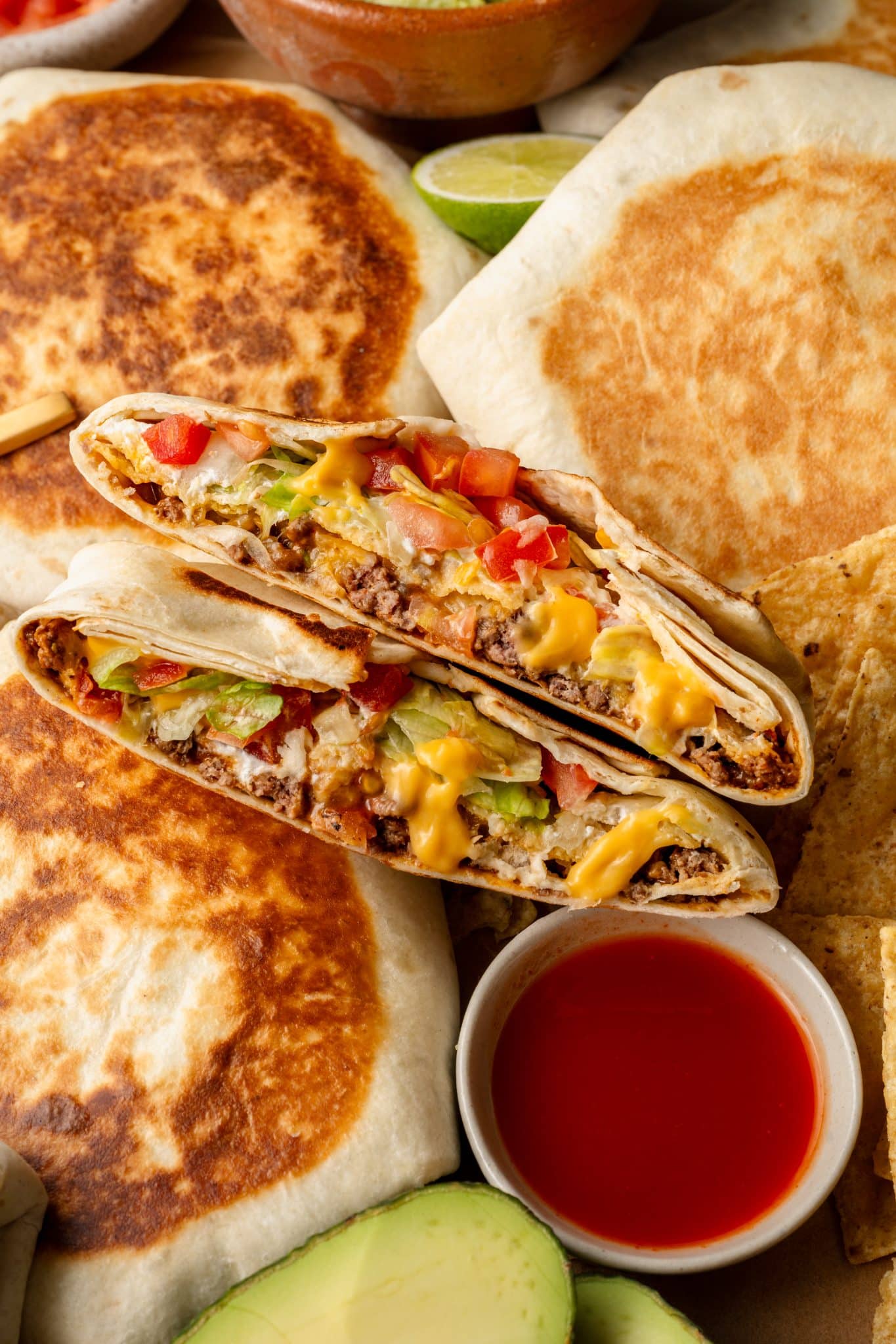 Close-up of a halved Crunchwrap Supreme stacked on top of golden-brown toasted Crunchwraps. The layers of seasoned ground beef, melted nacho cheese, crispy tostada shell, shredded lettuce, diced tomatoes, and sour cream are visible inside the soft flour tortilla. Surrounding elements include a bowl of red sauce, fresh avocado slices, tortilla chips, and lime wedges, creating a vibrant and appetizing presentation.