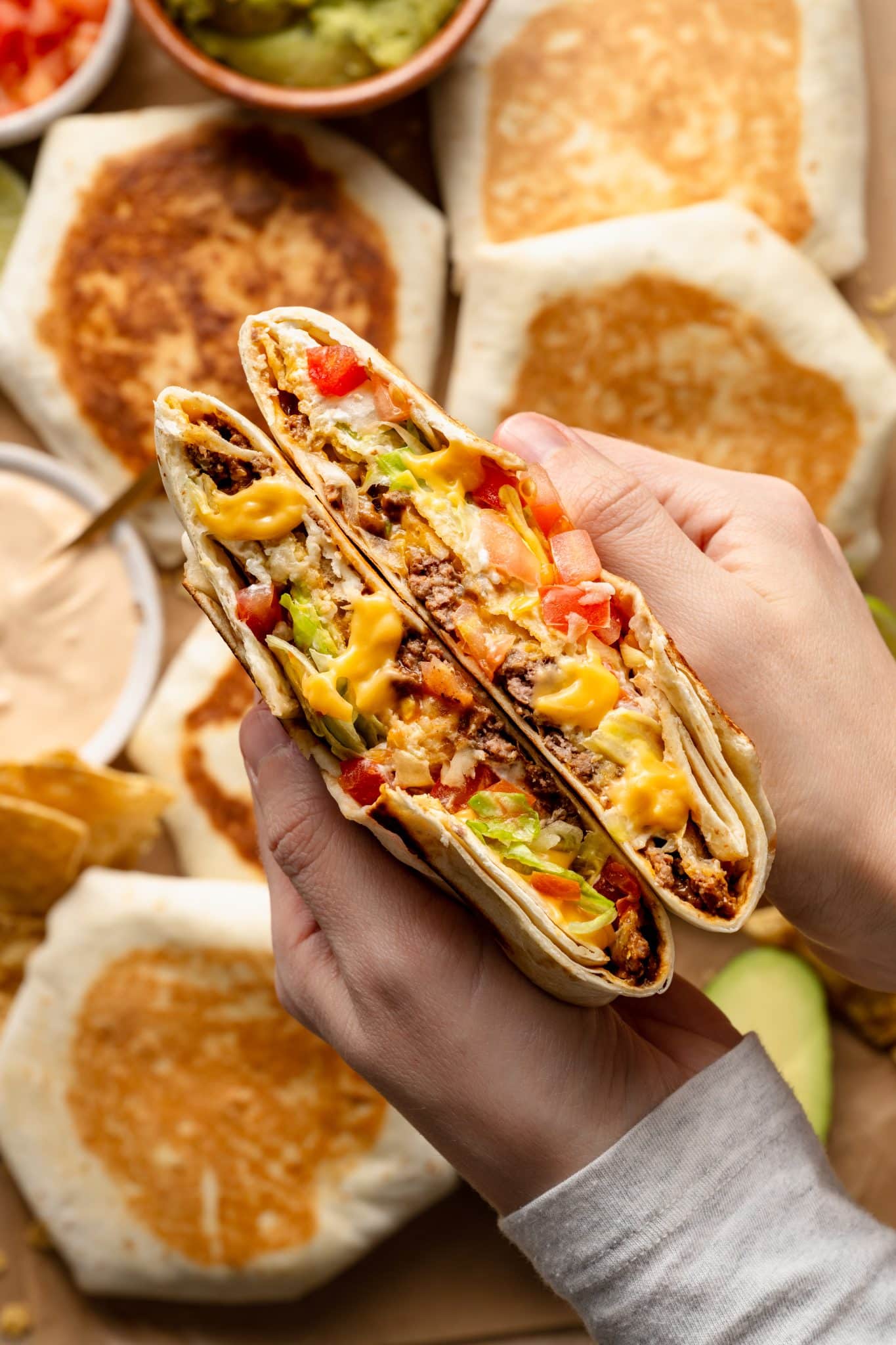 Close-up of hands holding a freshly cut Crunchwrap Supreme, revealing layers of seasoned ground beef, melted nacho cheese, crispy tostada shell, shredded lettuce, diced tomatoes, and sour cream inside a toasted flour tortilla. In the background, several whole Crunchwraps, bowls of guacamole, sauce, and tortilla chips are visible, adding to the appetizing presentation.