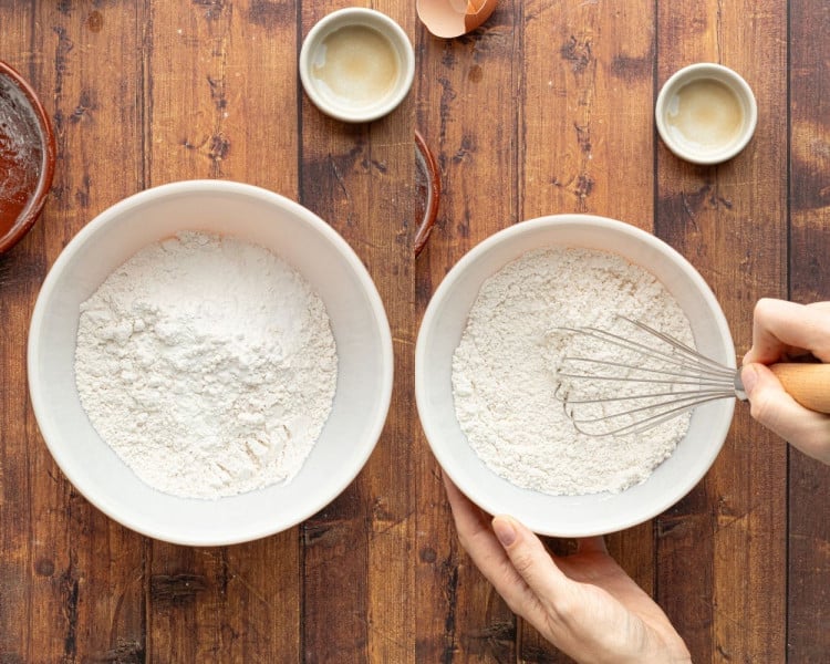 Two white mixing bowls with flour on a wooden surface. A person is whisking the flour in one of the bowls, while eggshells and small dishes are scattered around.