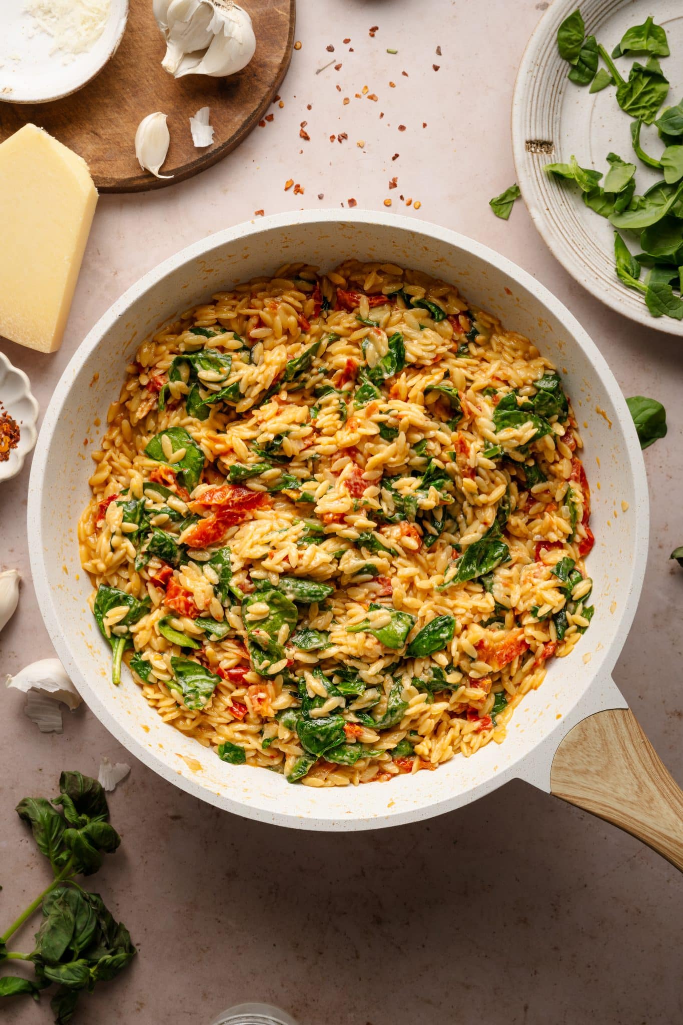 A creamy sun-dried tomato and spinach orzo dish in a white skillet, with a wooden spoon stirring the pasta. The dish is surrounded by fresh ingredients like garlic, Parmesan cheese, and red pepper flakes on a neutral-toned countertop.