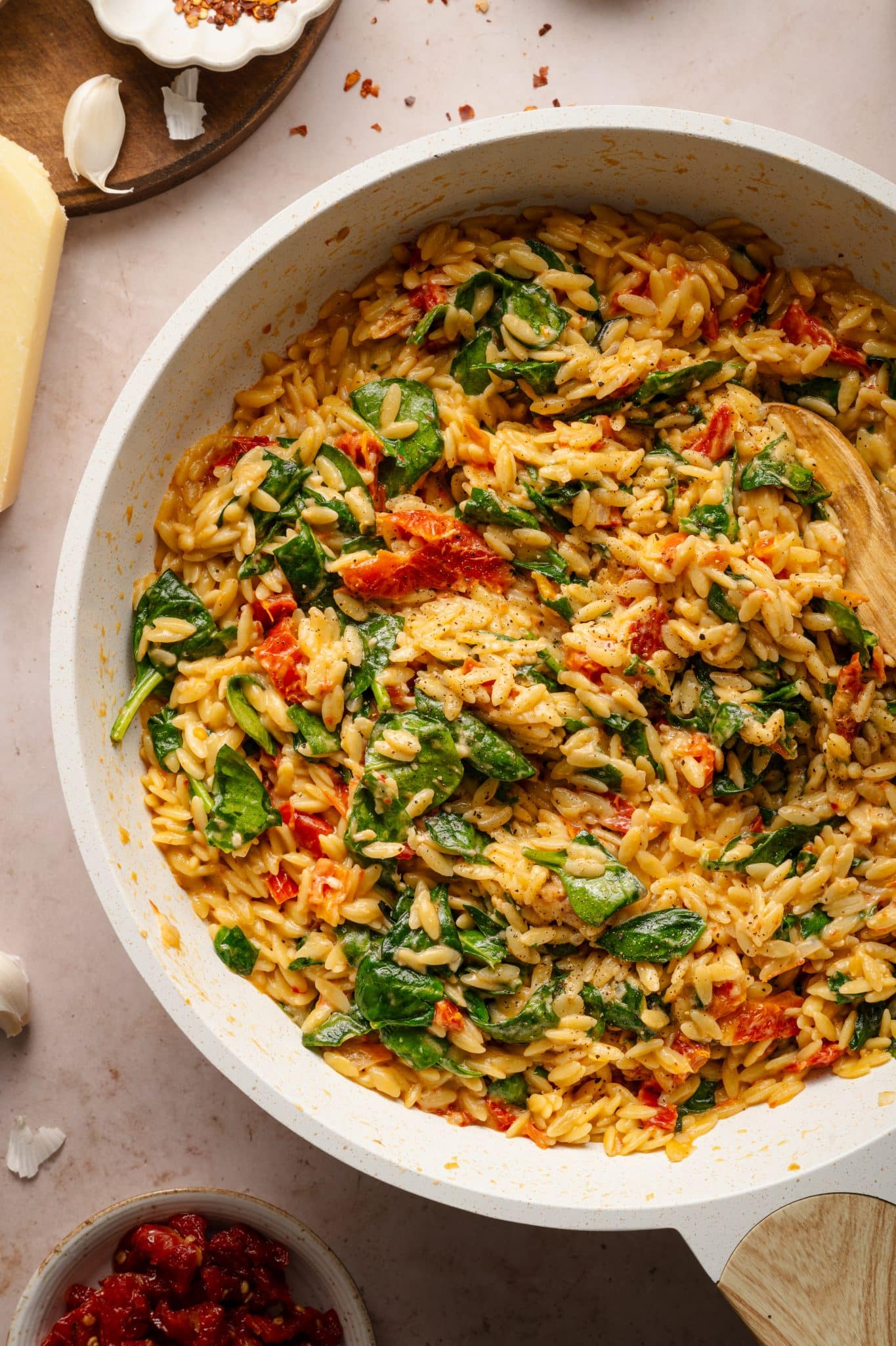 A creamy sun-dried tomato and spinach orzo dish in a white skillet, garnished with fresh spinach and sun-dried tomatoes. The orzo is coated in a rich, velvety sauce, with flecks of black pepper and Parmesan cheese. A wooden spoon rests in the pan, ready for serving, while surrounding ingredients like garlic, Parmesan, and red pepper flakes add to the rustic kitchen scene.