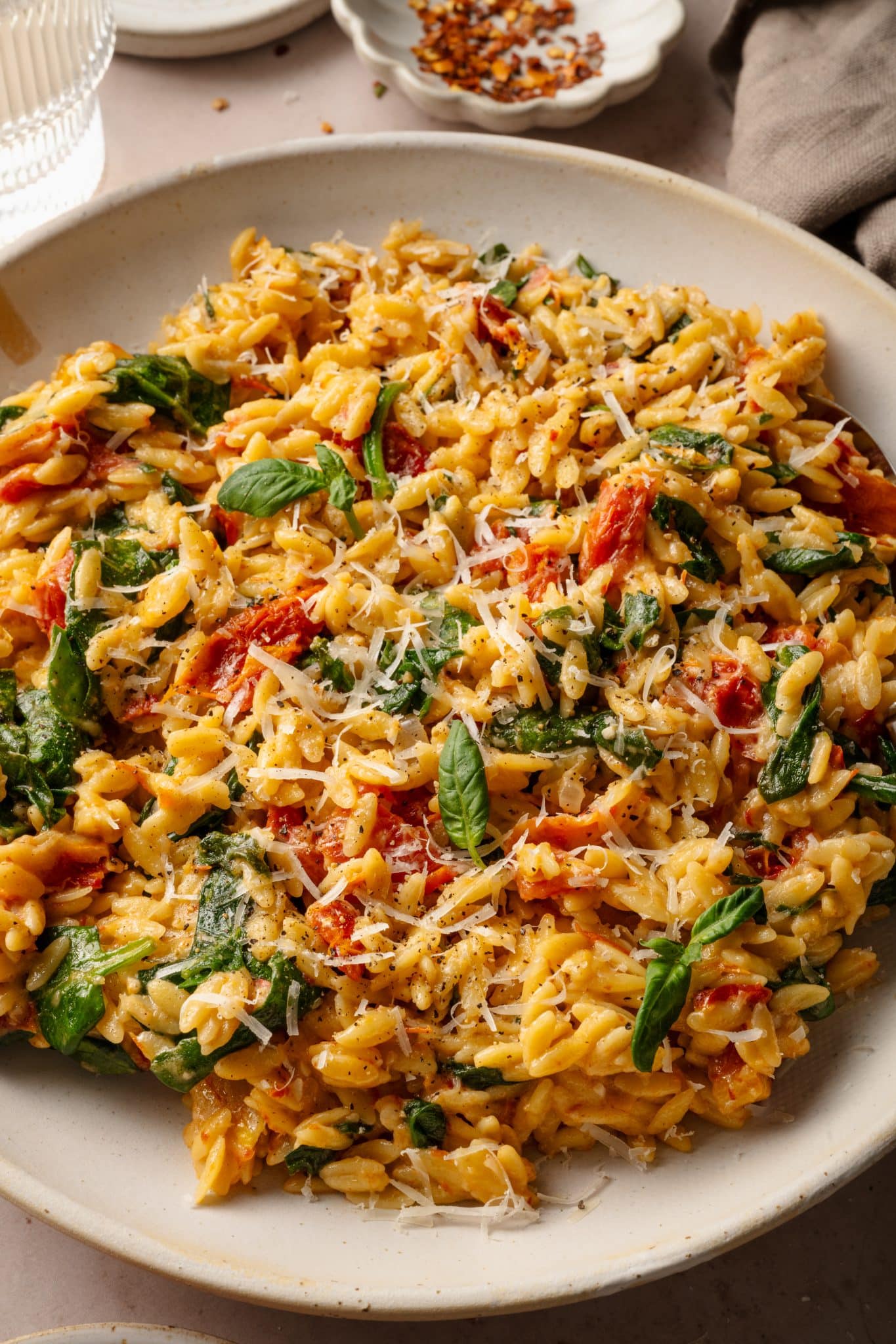 A plated serving of creamy sun-dried tomato and spinach orzo, garnished with fresh basil and grated Parmesan cheese. The dish is set on a neutral plate with a rustic beige napkin and a small bowl of red pepper flakes in the background.