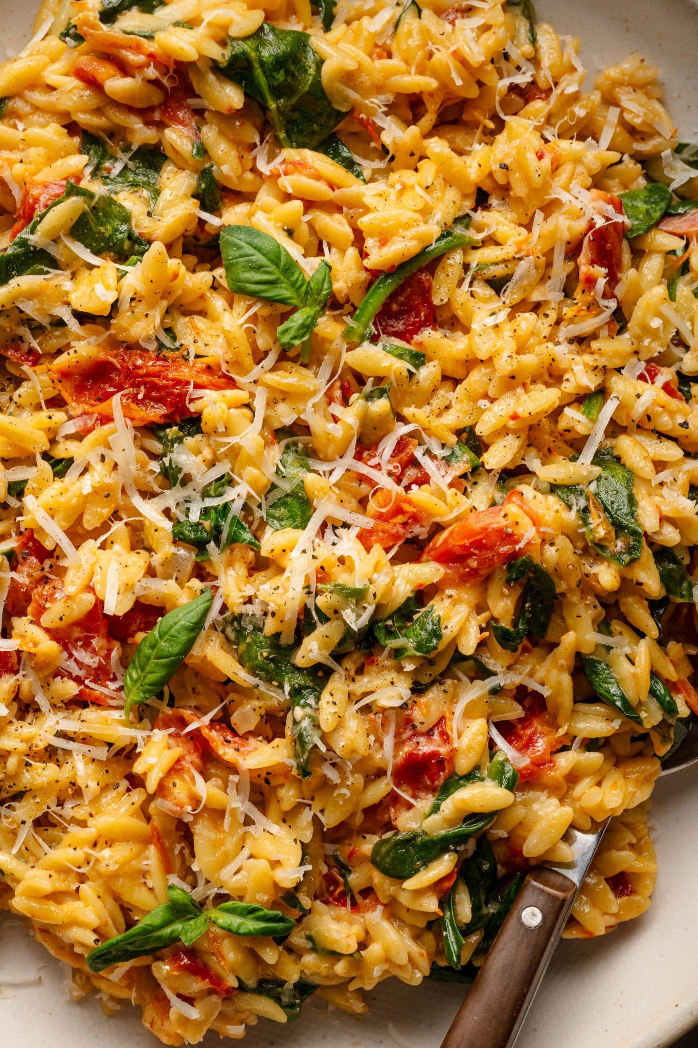 A close-up shot of creamy sun-dried tomato and spinach orzo, garnished with fresh basil leaves and freshly grated Parmesan cheese. The vibrant red sun-dried tomatoes, wilted green spinach, and golden orzo are coated in a rich, creamy sauce, creating a visually appetizing and flavorful dish. A fork rests on the plate, ready for serving.